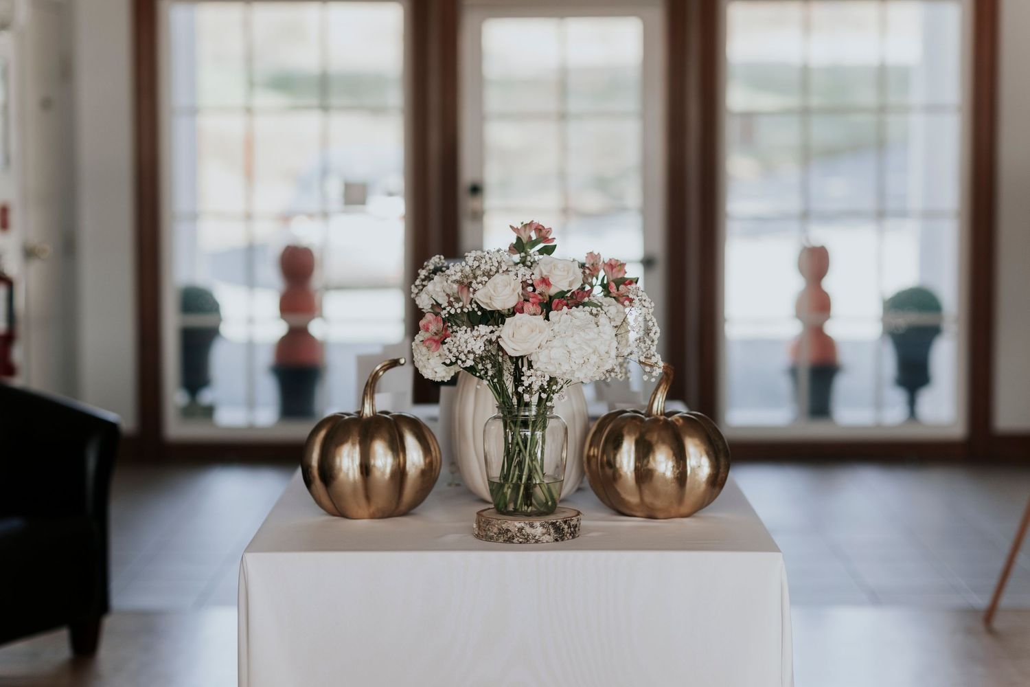 Gold metallic pumpkins flank a delicate floral centerpiece on a white table near windows.