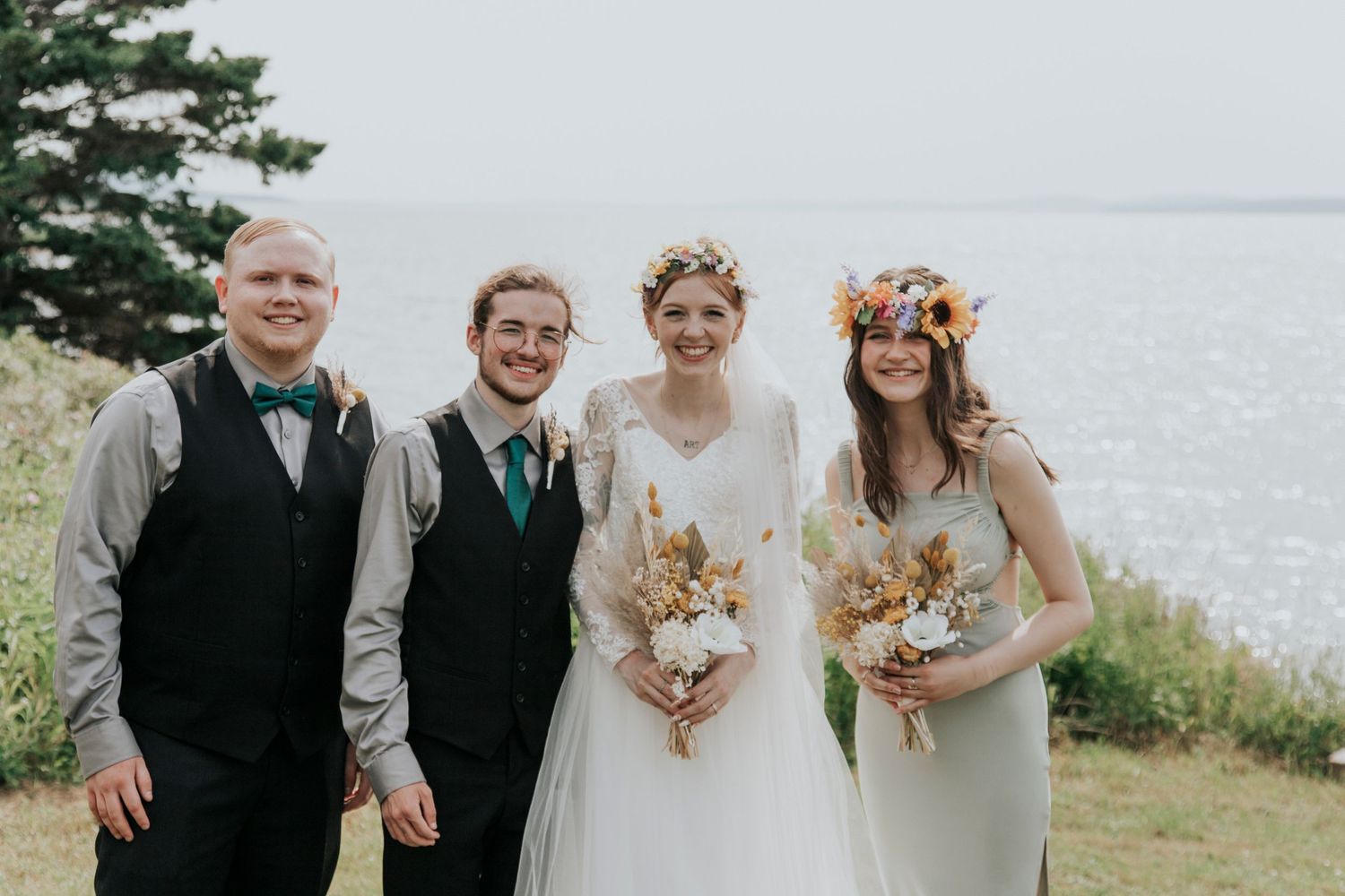 Wedding party stands on coastal hillside overlooking Bras d'Or while holding bouquets and wearing formal attire.