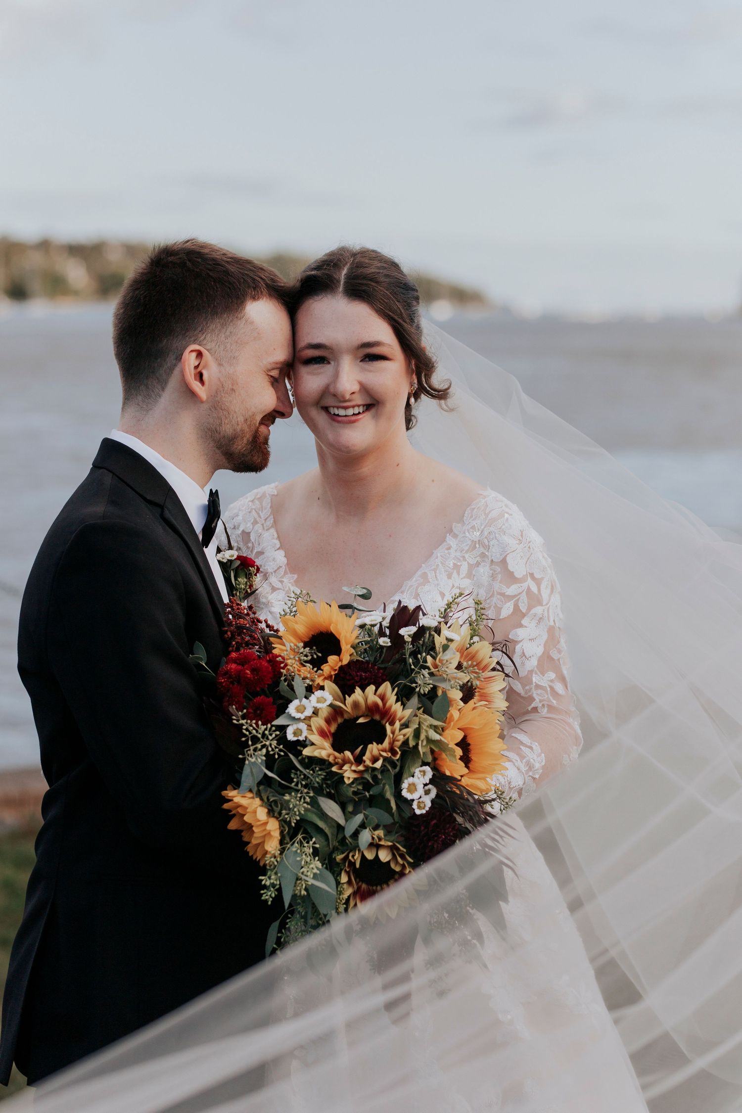 Bride holds sunflower bouquet as veil blows in wind beside groom near waterfront.