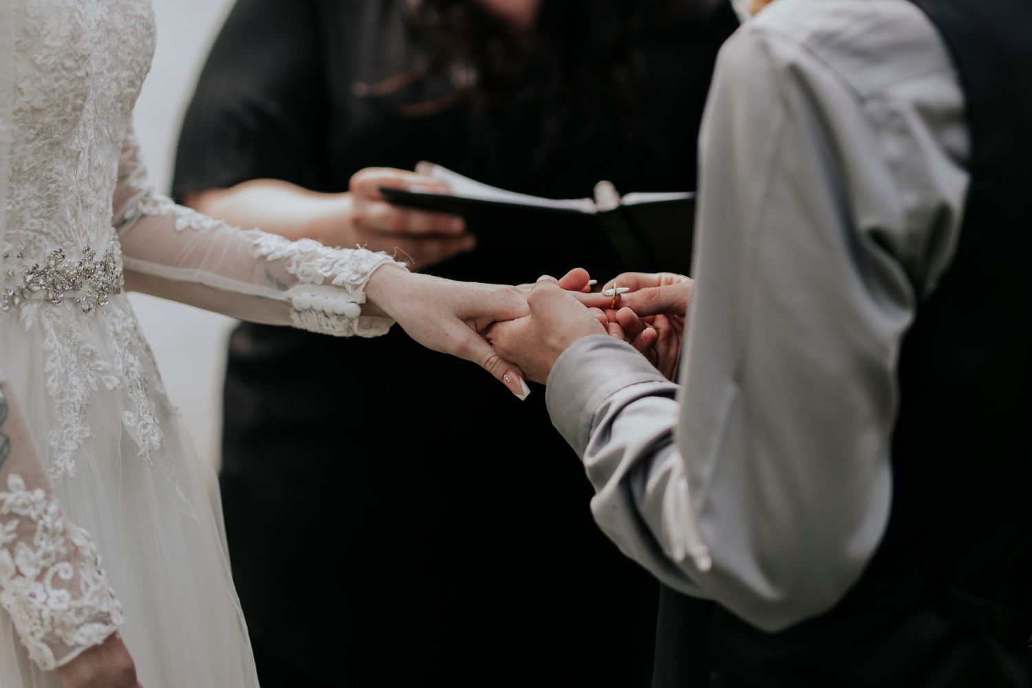 Close-up shot of exchanging wedding rings during ceremony with lace sleeve detail and officiant in frame.