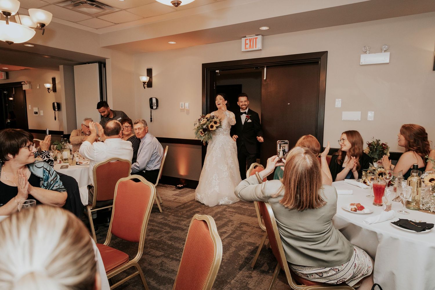 Wedding reception scene with guests seated at round tables while couple enters the room.