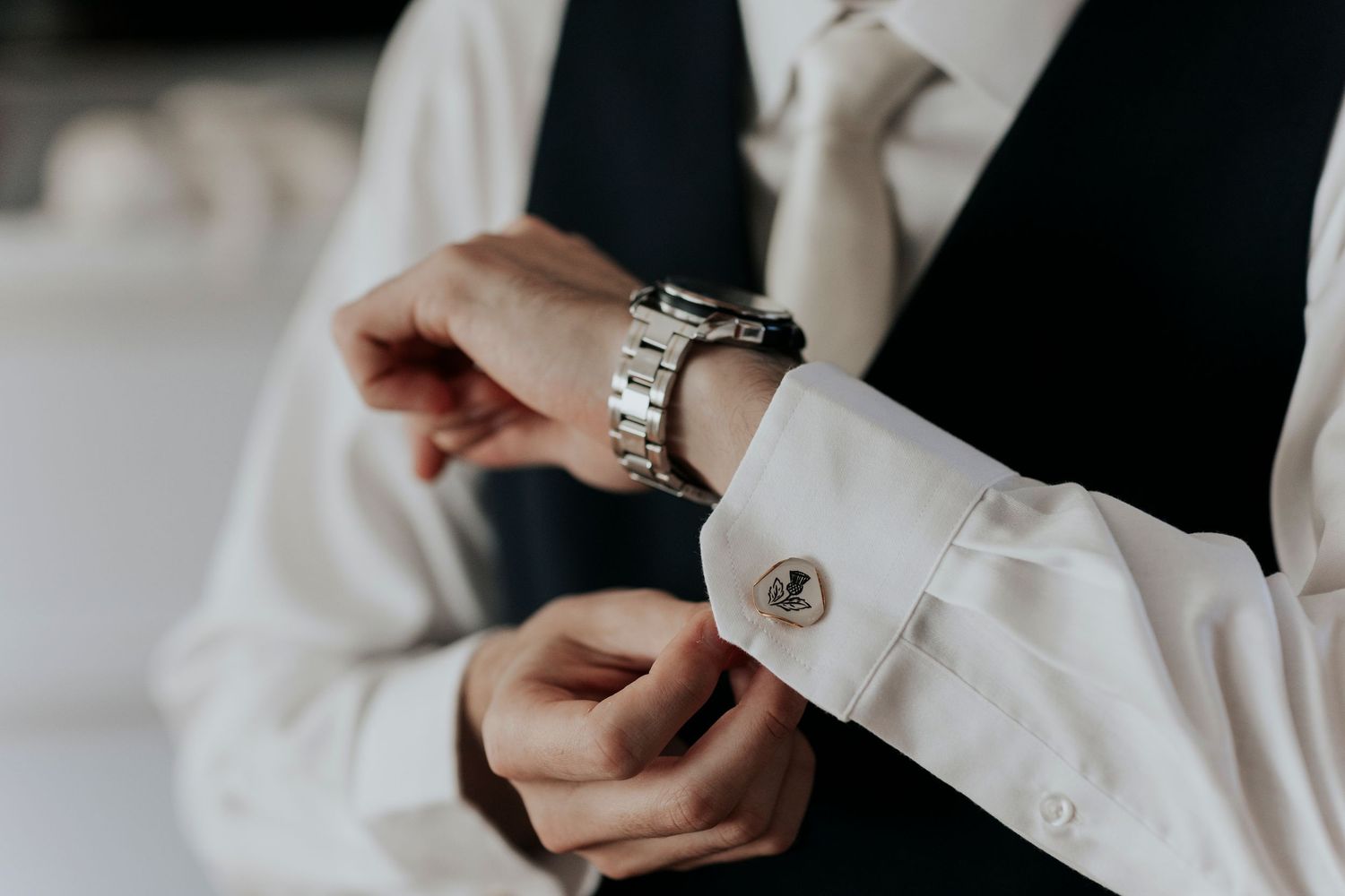 Close-up of hands adjusting a silver watch and cufflink on a white dress shirt sleeve with black suit vest.