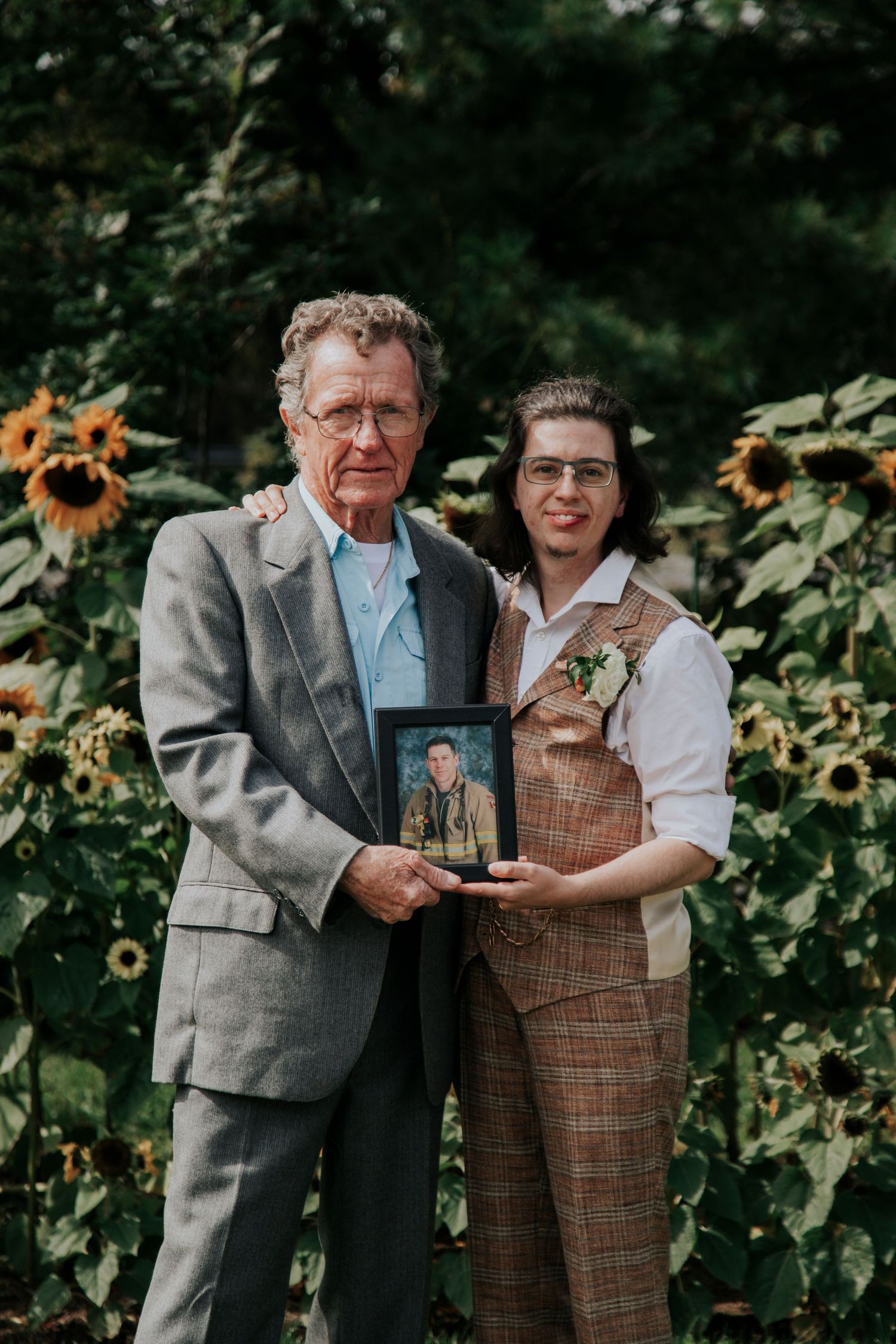 Two people stand together against a garden backdrop with sunflowers while holding a framed photograph.