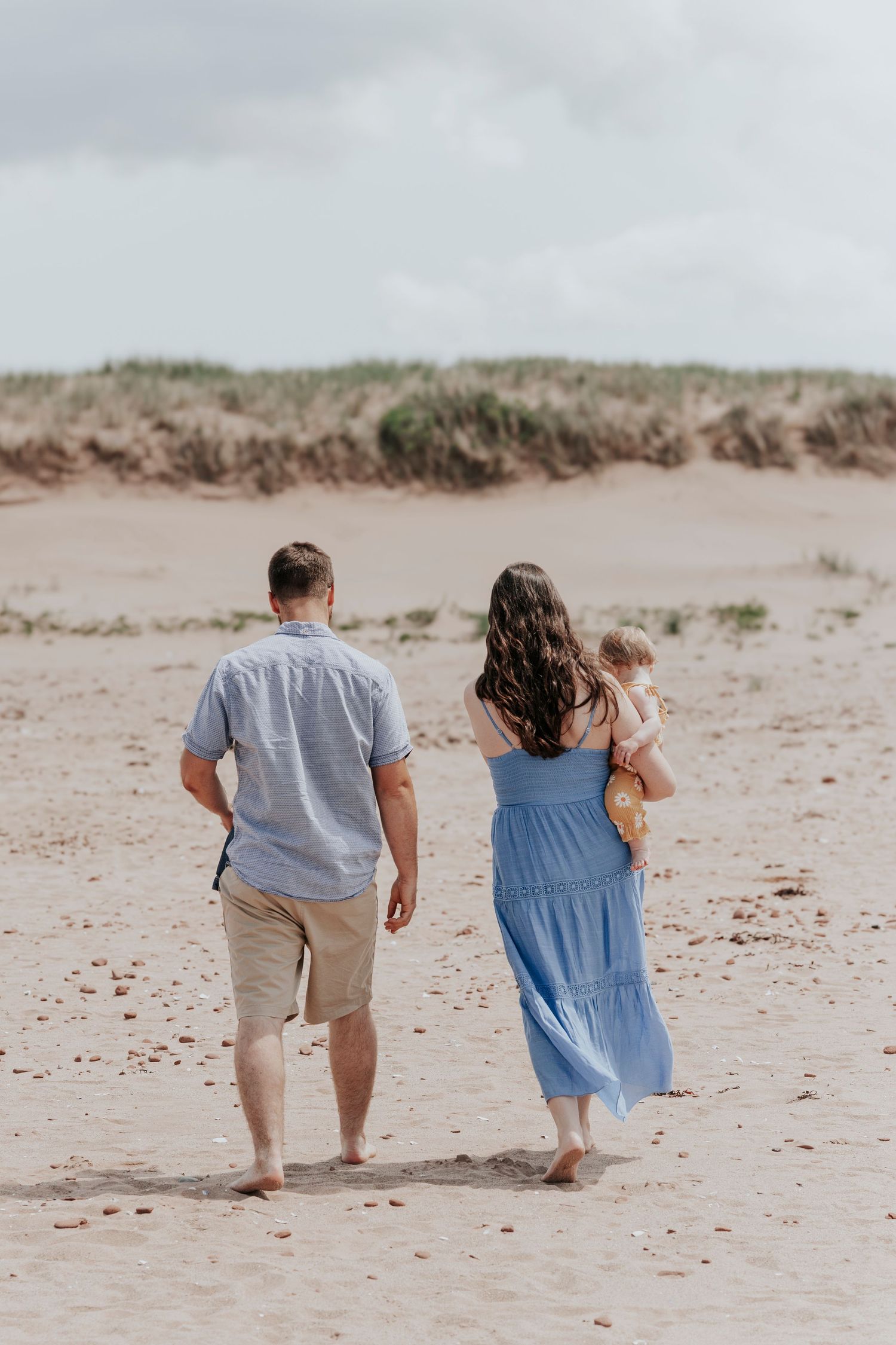 Family walking on beach, mother carrying baby in daisy outfit.