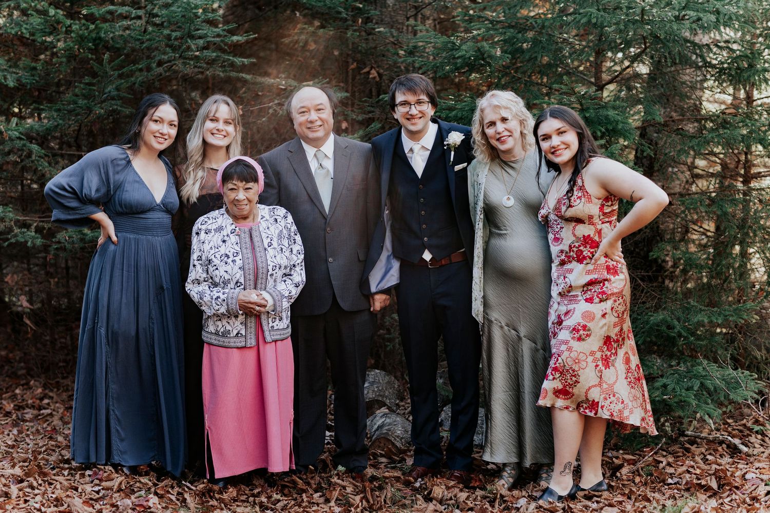 A group of seven people in formal attire pose together outdoors against a wooded forest backdrop.