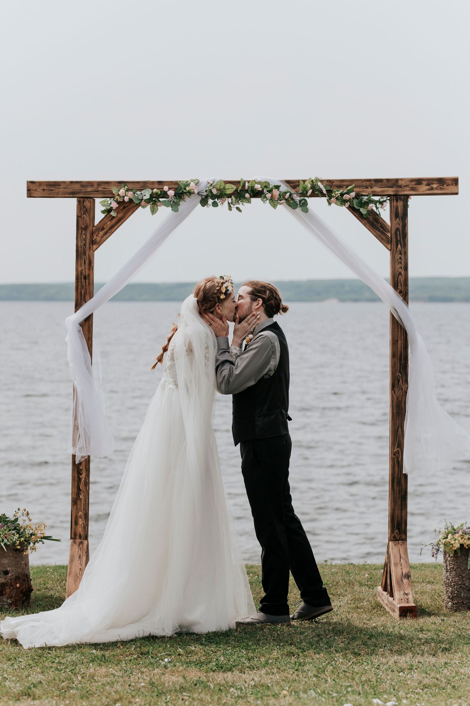 A couple shares their first kiss under a wooden wedding arch decorated with white fabric and greenery by the waterfront.