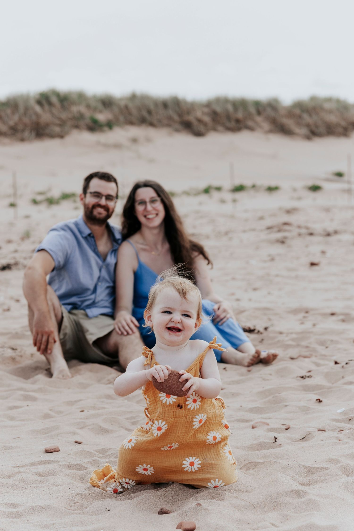 Happy baby in a daisy romper sits on sandy beach while parents relax together in the background.