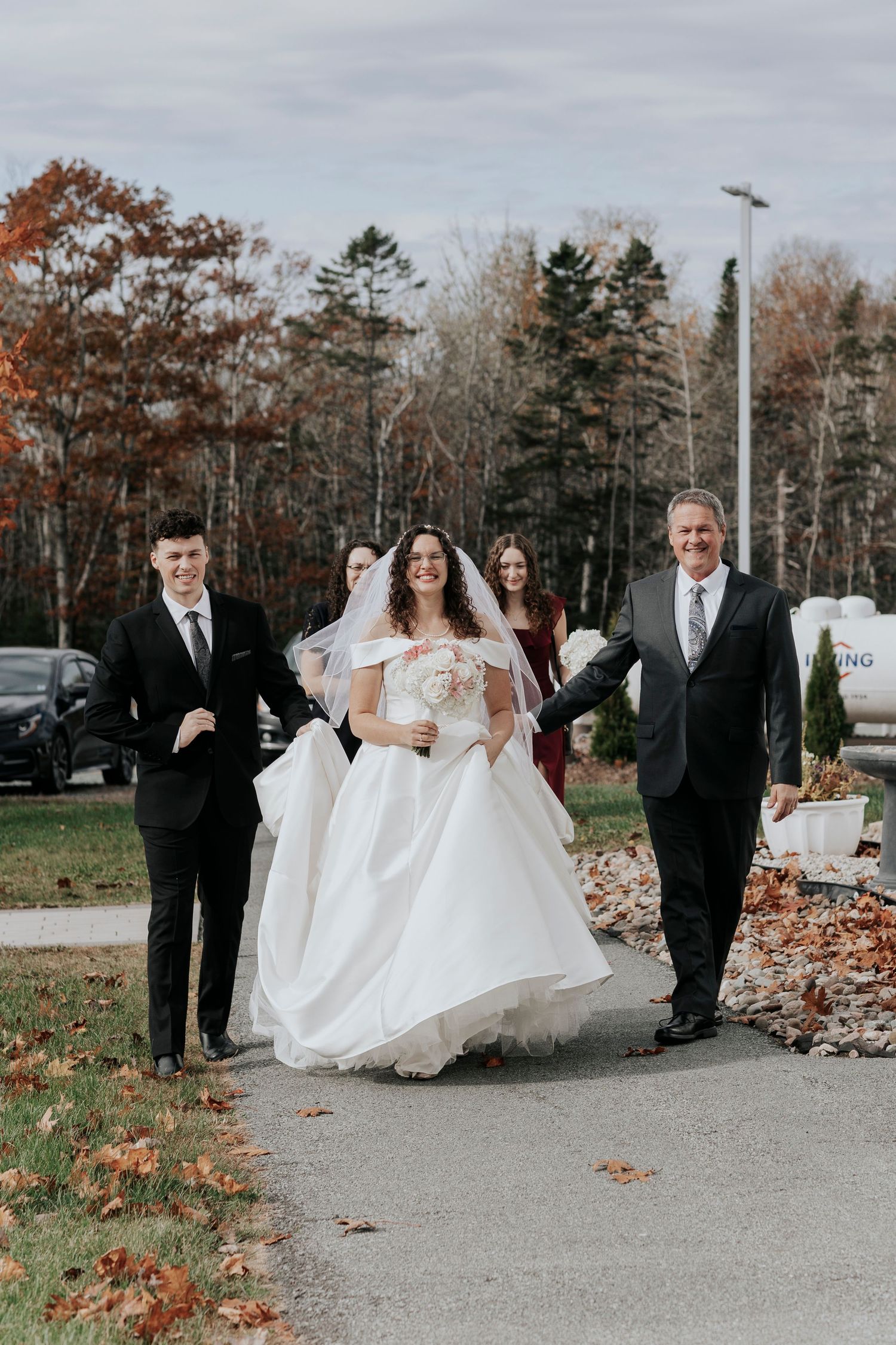 A joyful wedding party walks along a path lined with autumn trees on an overcast day.