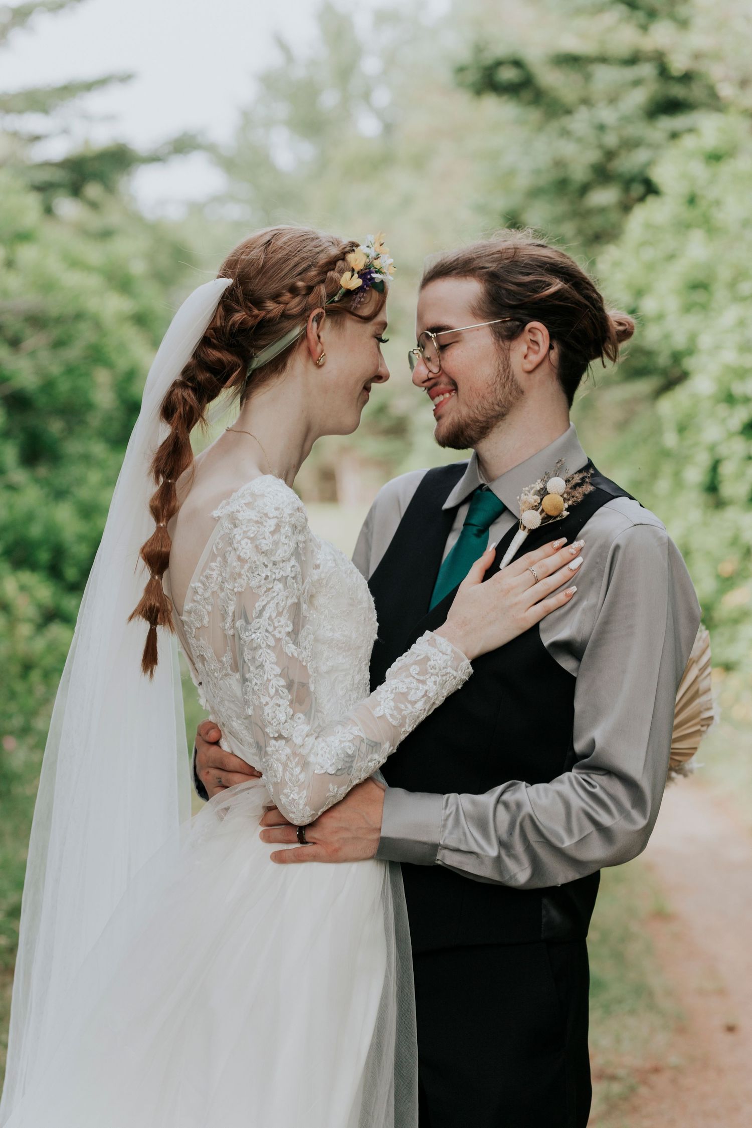 A romantic forest wedding portrait shows a couple in white lace and grey vest sharing an intimate moment outdoors.