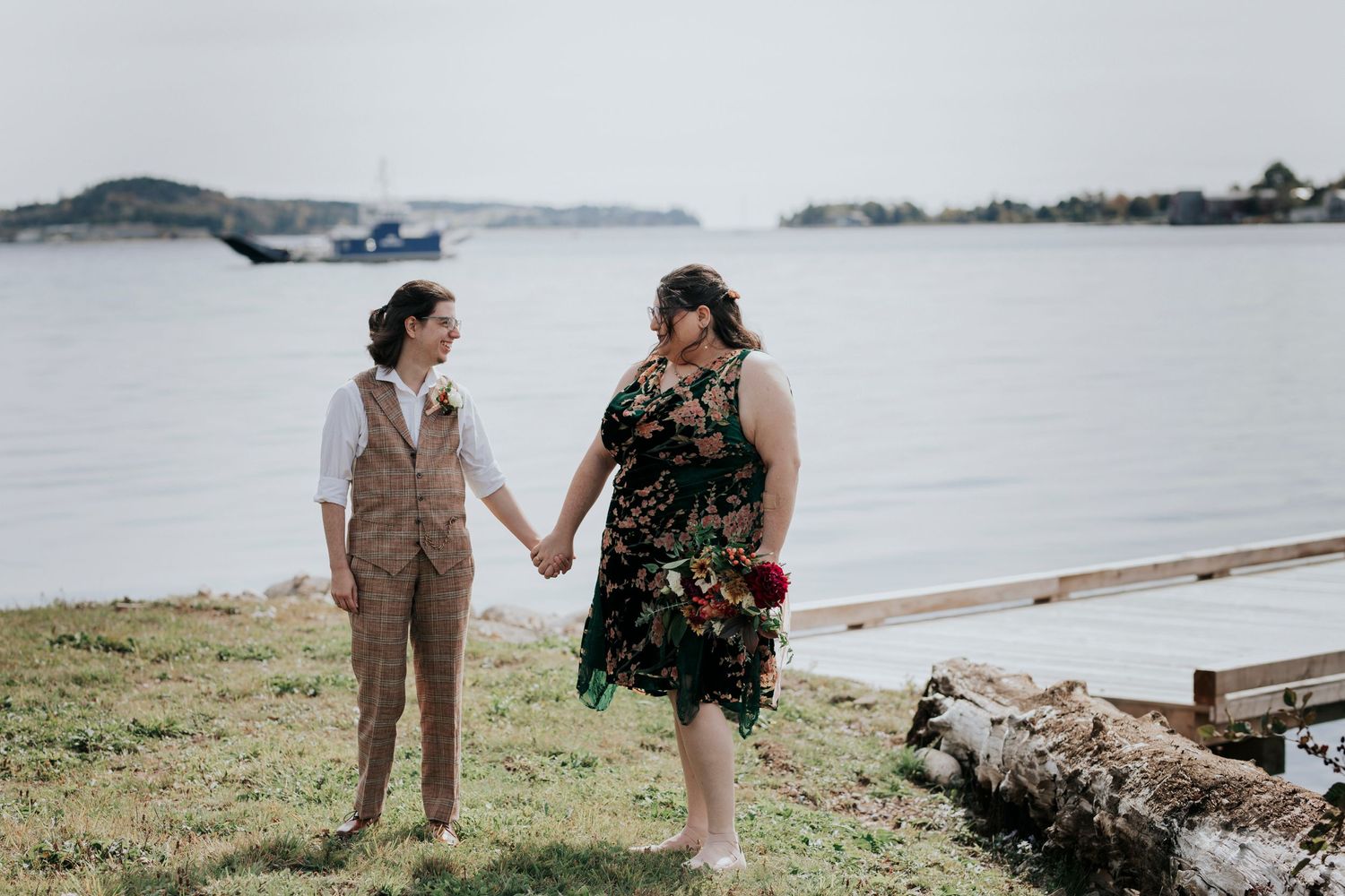 A wedding couple holds hands while standing on a grassy shore overlooking a misty lake and distant islands.