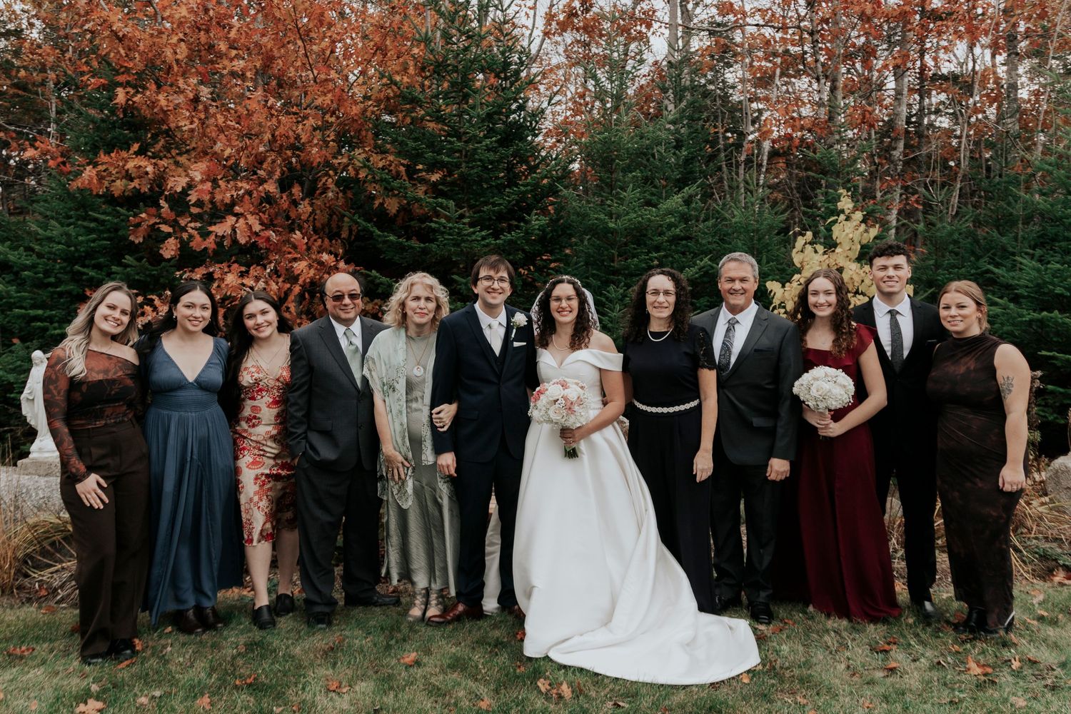 A wedding party stands together outdoors against a backdrop of autumn trees.