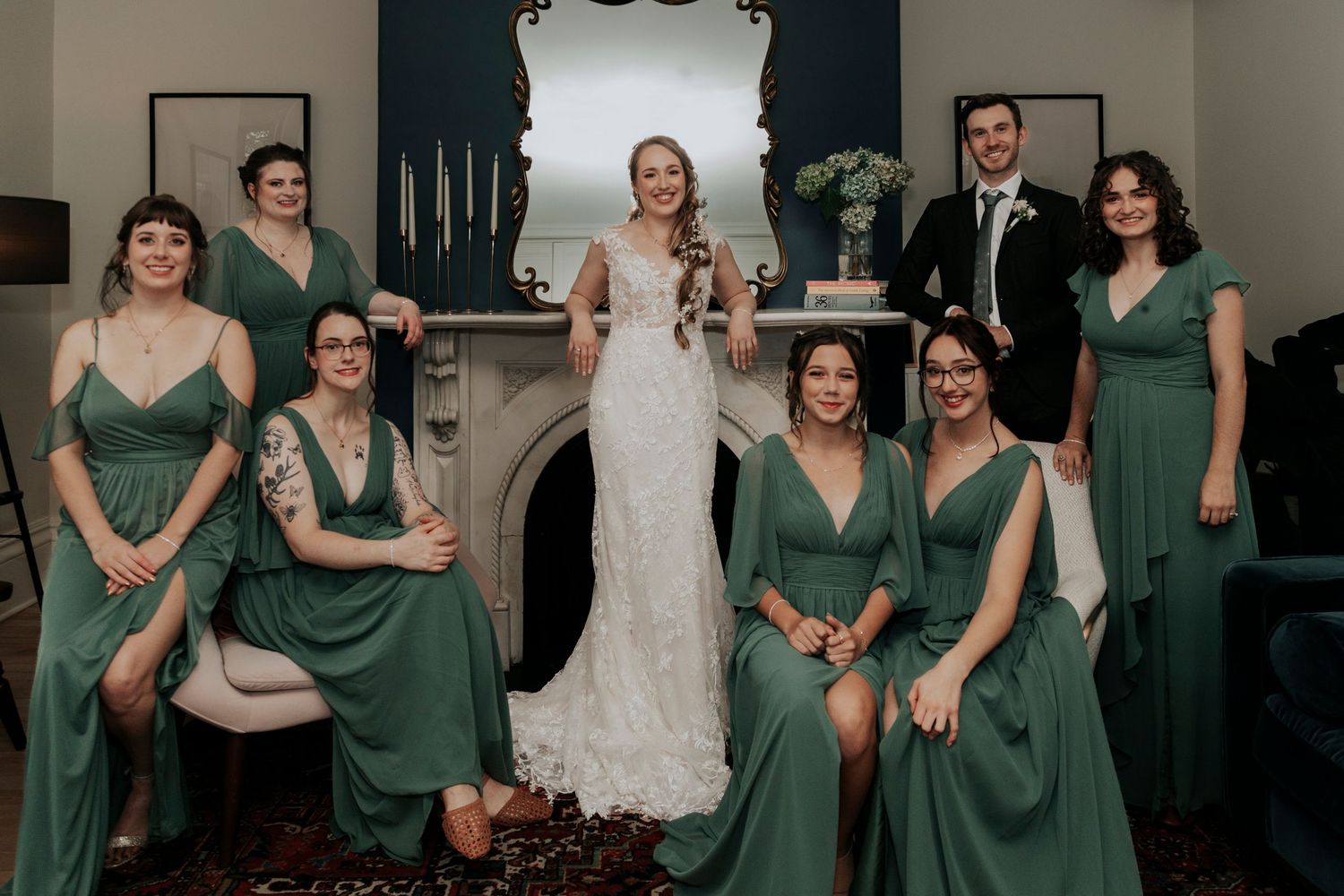 Bride poses with bridesmaids in elegant green dresses near a fireplace at wedding reception.