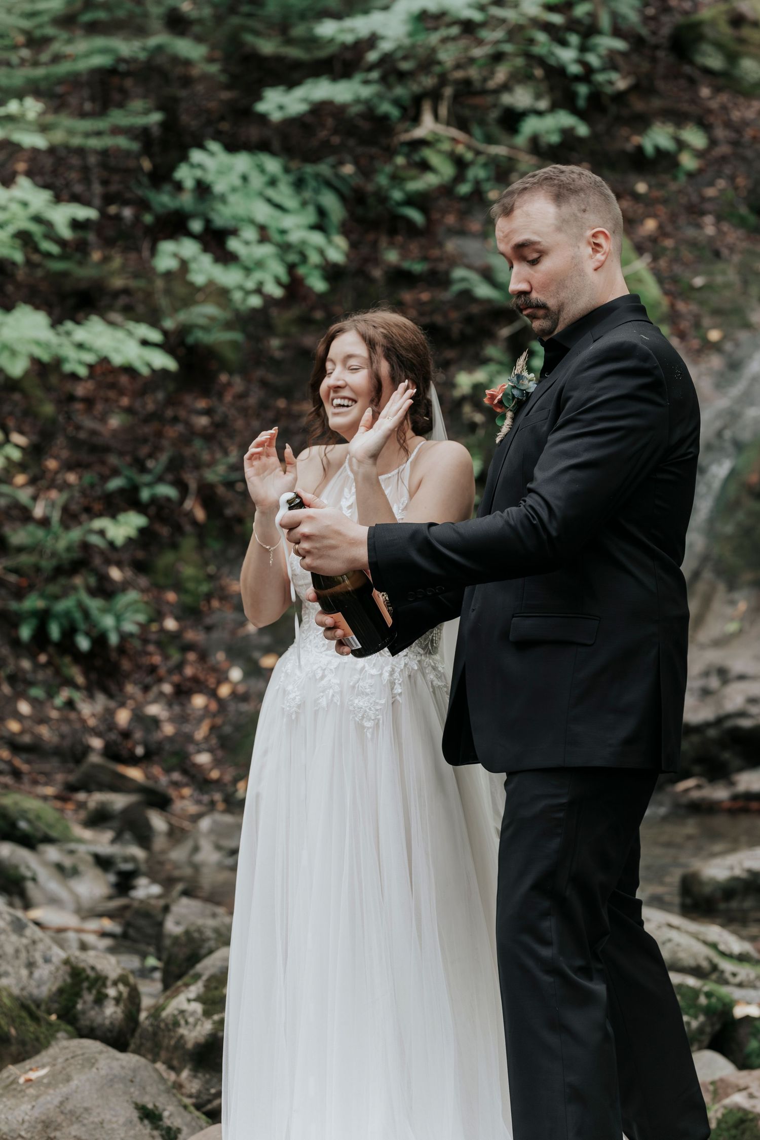 Joyful bride laughs as groom pops champagne bottle to celebrate their elopement in a forest setting.