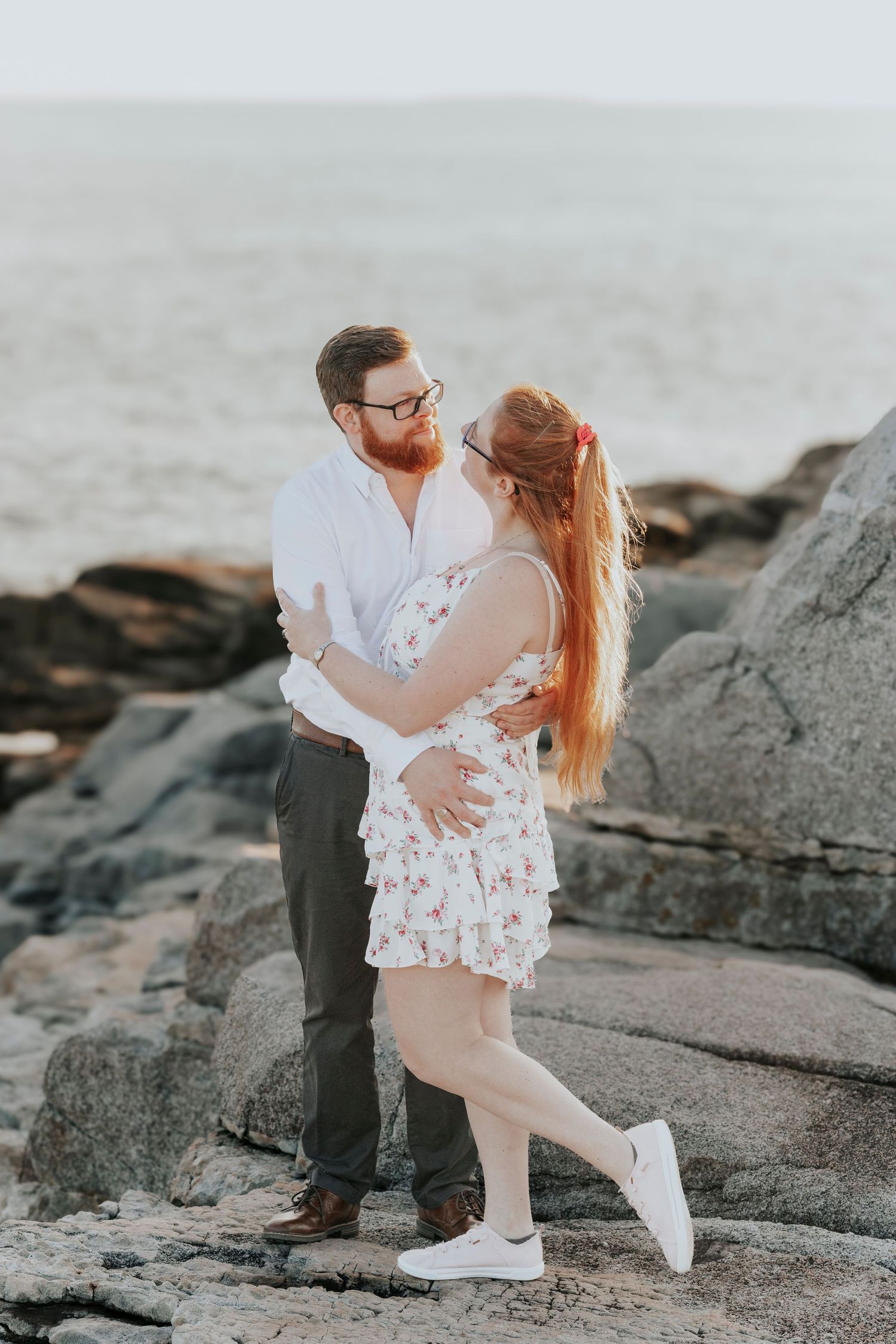 Couple in white outfits embracing romantically on rocky coastline with ocean waves and boulders behind them.