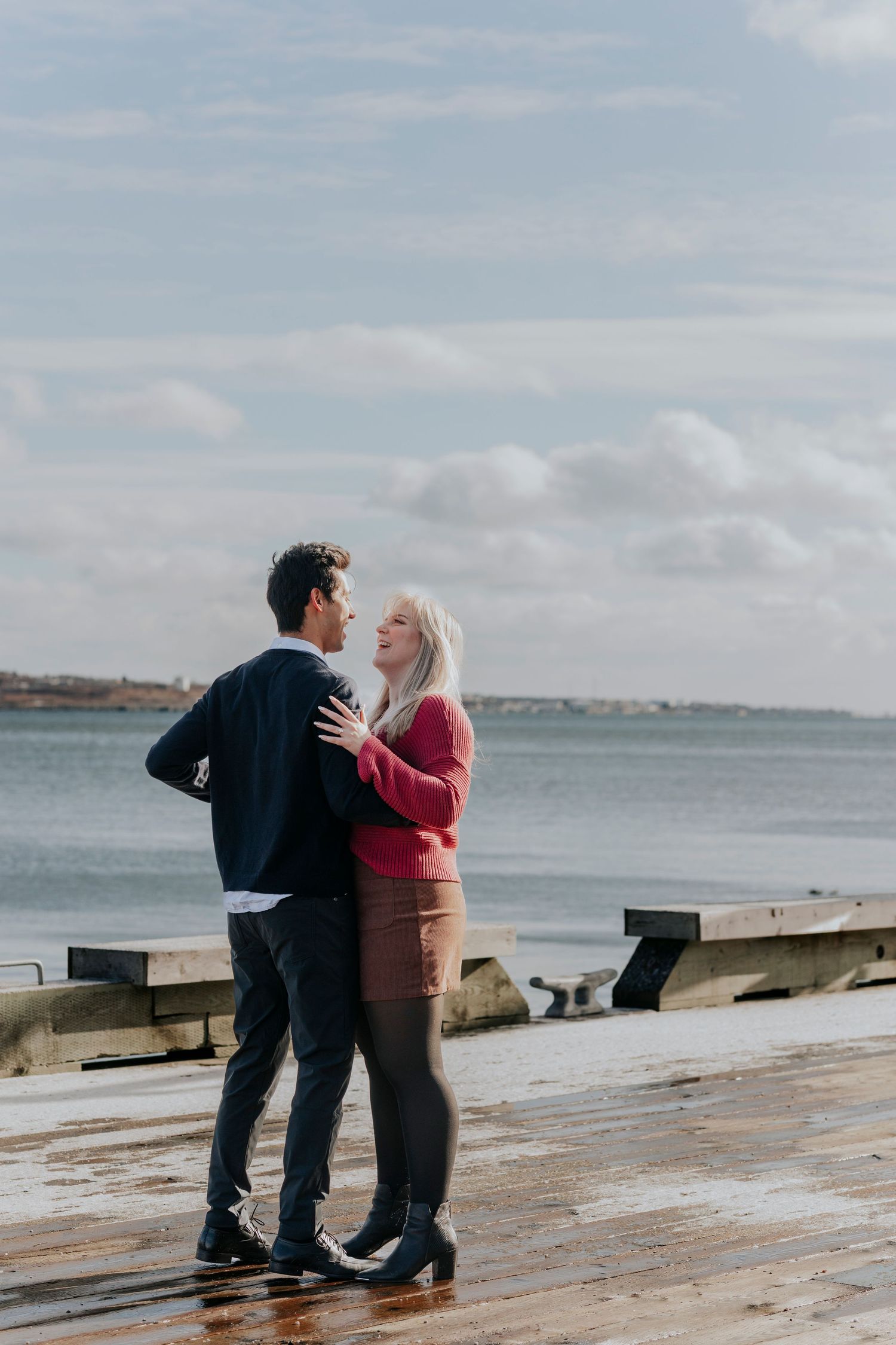 Couple embracing and gazing at each other on a wet waterfront pier with ocean views behind them.