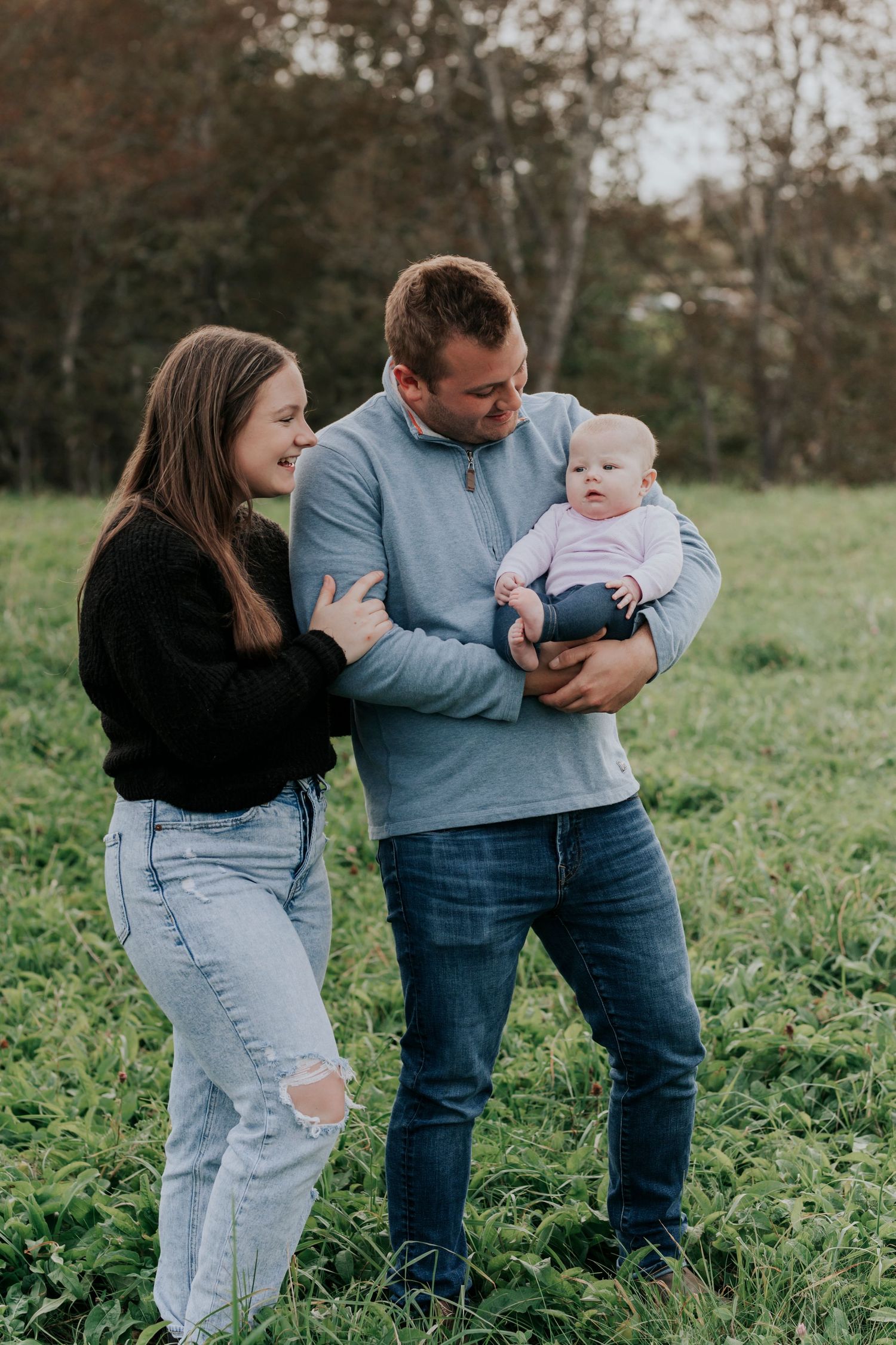 Young parents stand in a green field, smiling at their newborn baby during an outdoor family photo session.