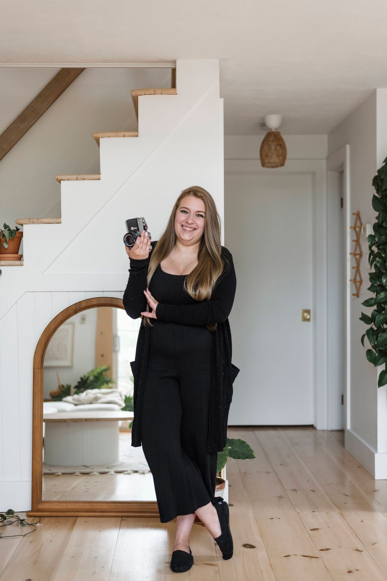 Smiling female photographer in all-black outfit holding camera inside modern home with staircase.