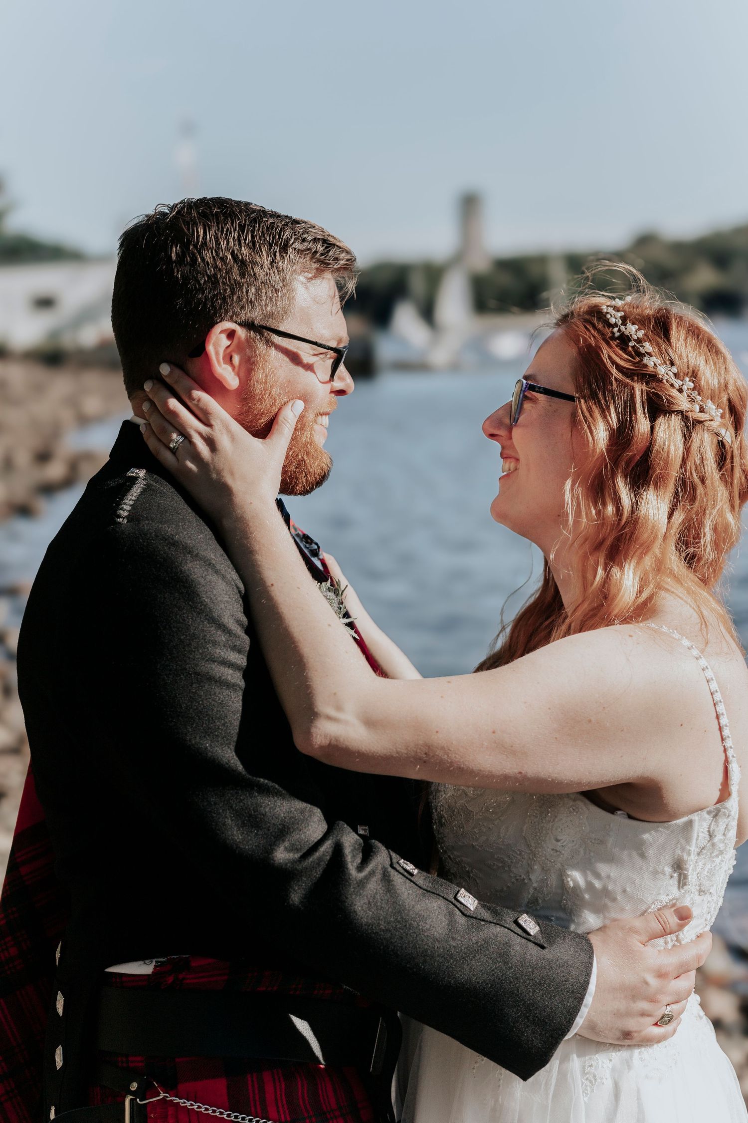 Bride and groom in tartan kilt sharing a tender moment by the waterfront on their wedding day.