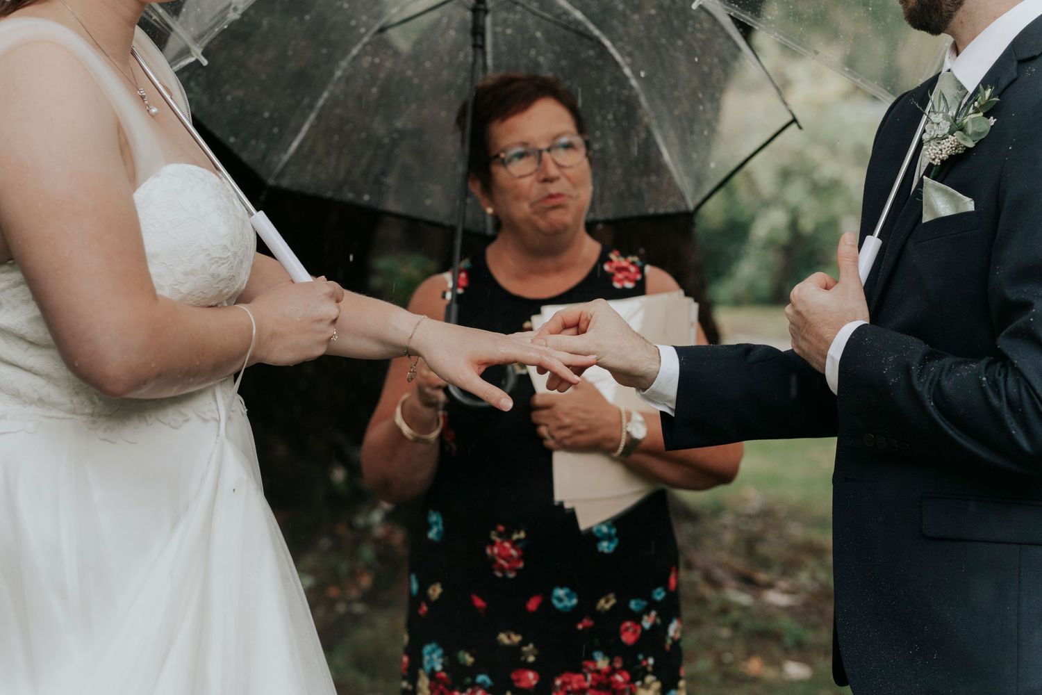 Officiant places wedding ring on bride's finger during an outdoor rainy ceremony under a clear umbrella.