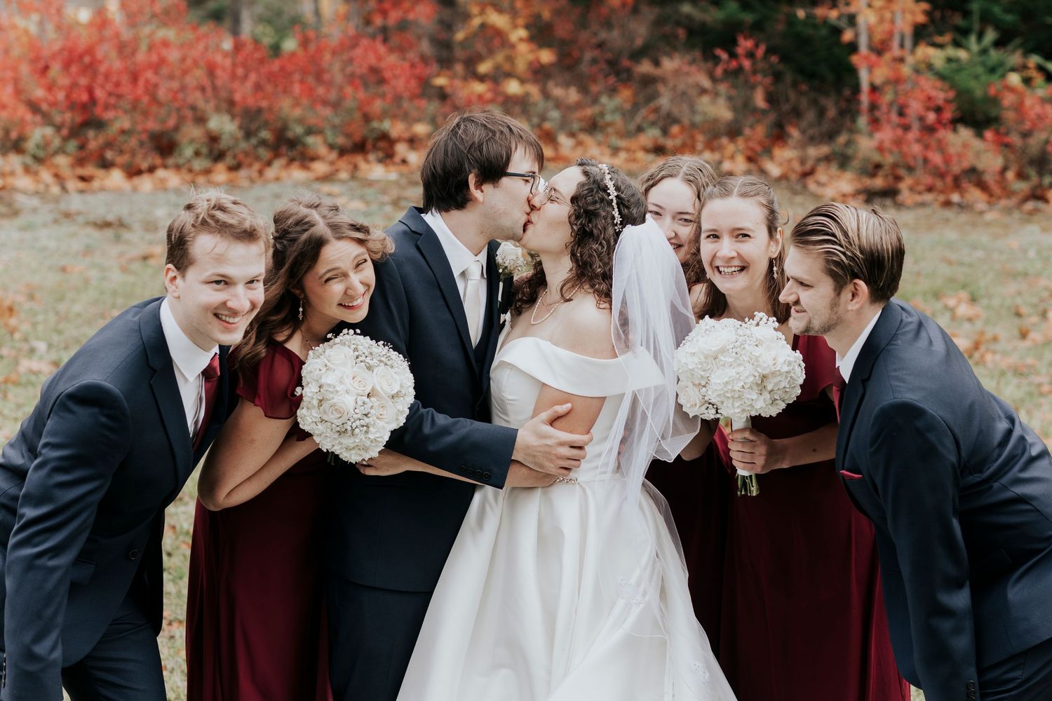 Wedding party in burgundy and navy attire surrounds kissing couple among autumn foliage.