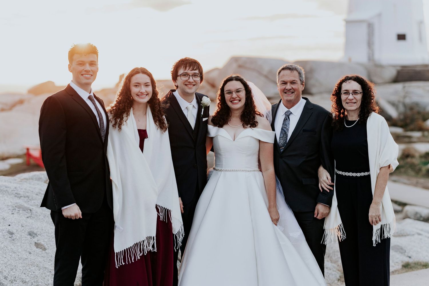 A bride in white dress poses with family members outdoors at sunset near coastal location.