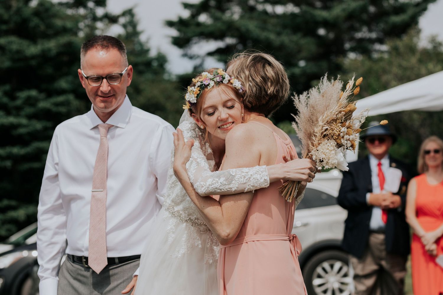 A bride in a white lace dress embraces her mom during an outdoor wedding celebration.