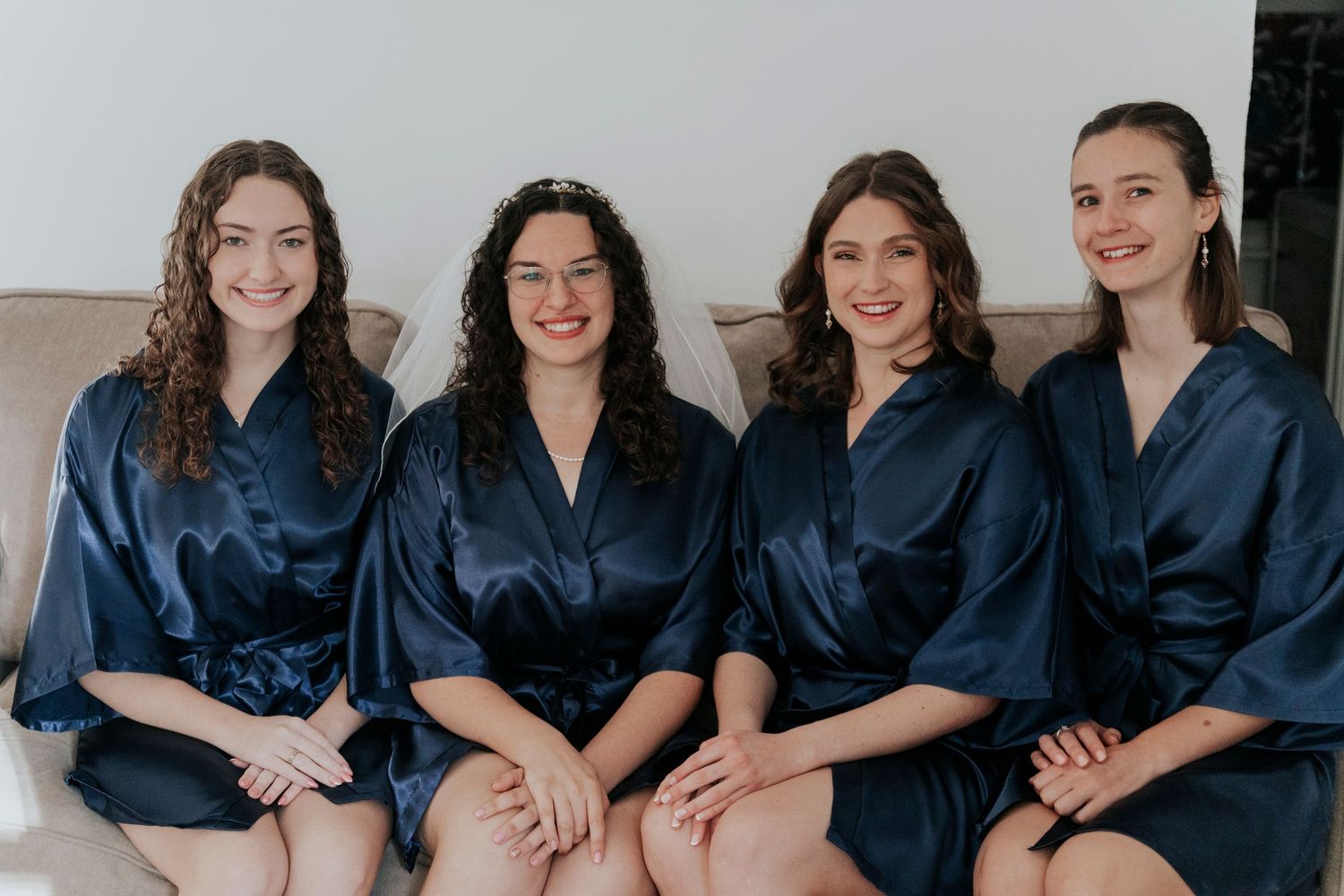 A group of four bridesmaids wearing matching navy blue silk robes sit together smiling while getting ready.