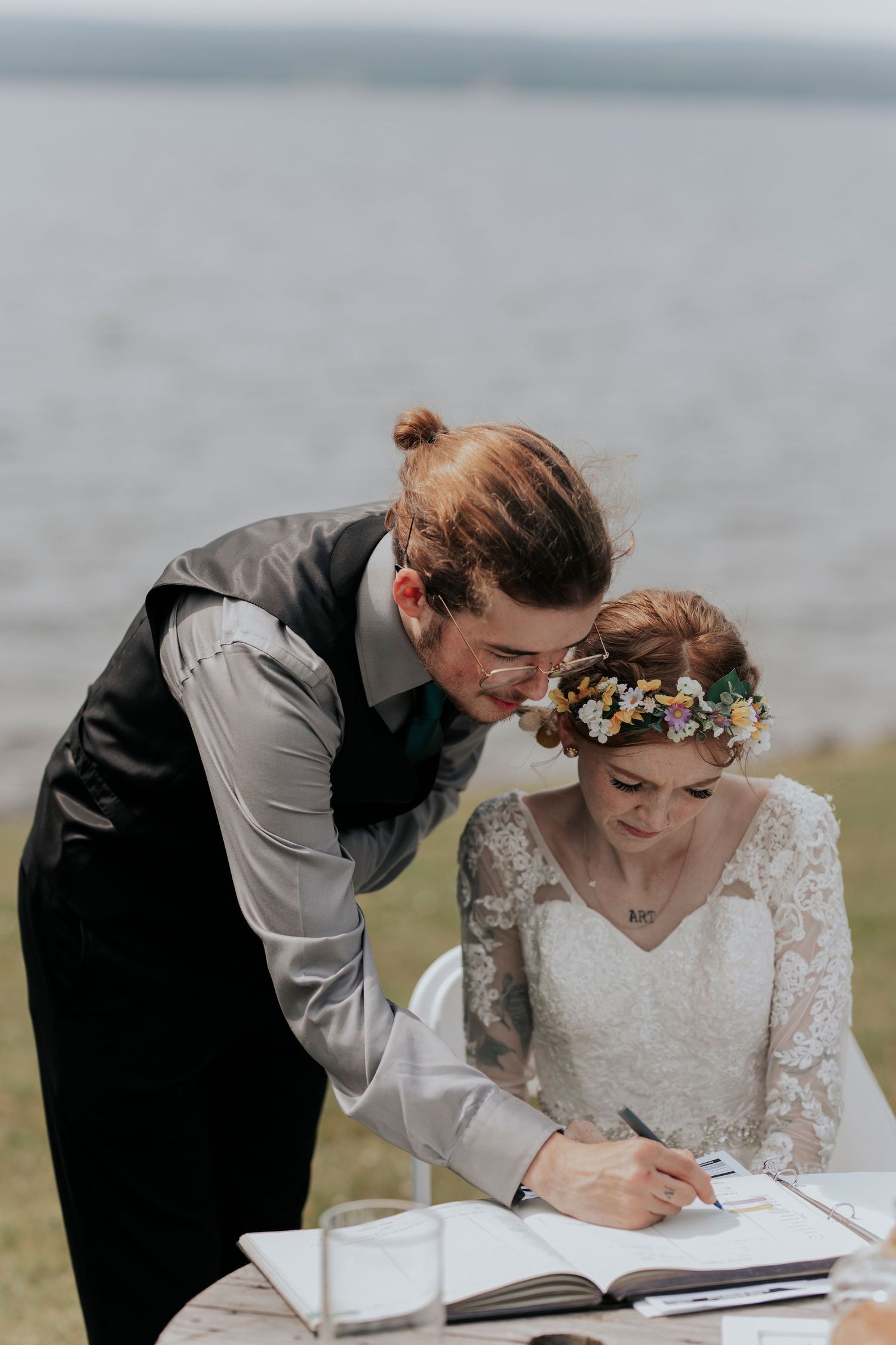 Couple signing wedding registry outdoors, bride wearing lace sleeves and floral crown near waterfront.