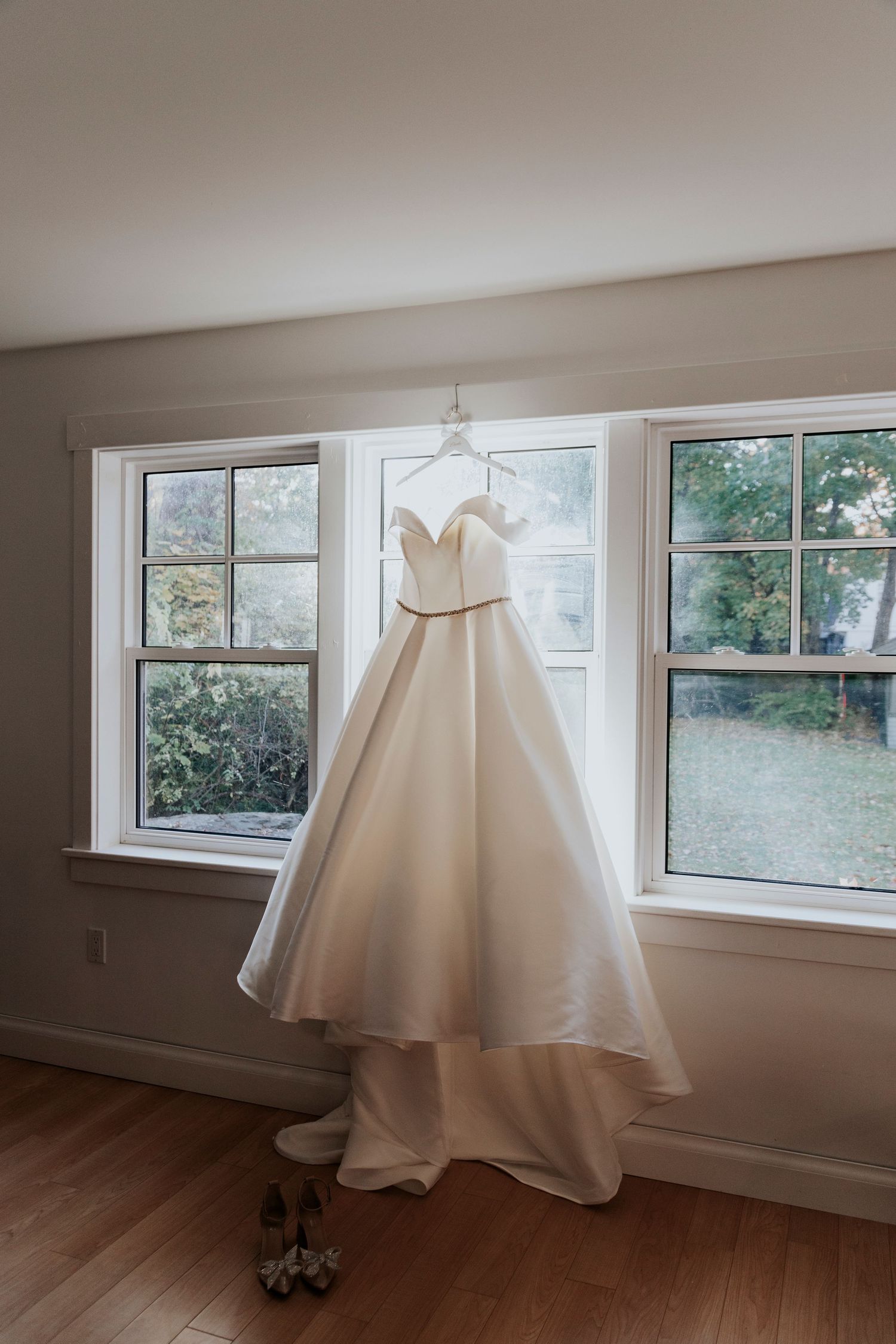 Elegant off-shoulder white wedding ball gown hanging in window with bridal shoes below.