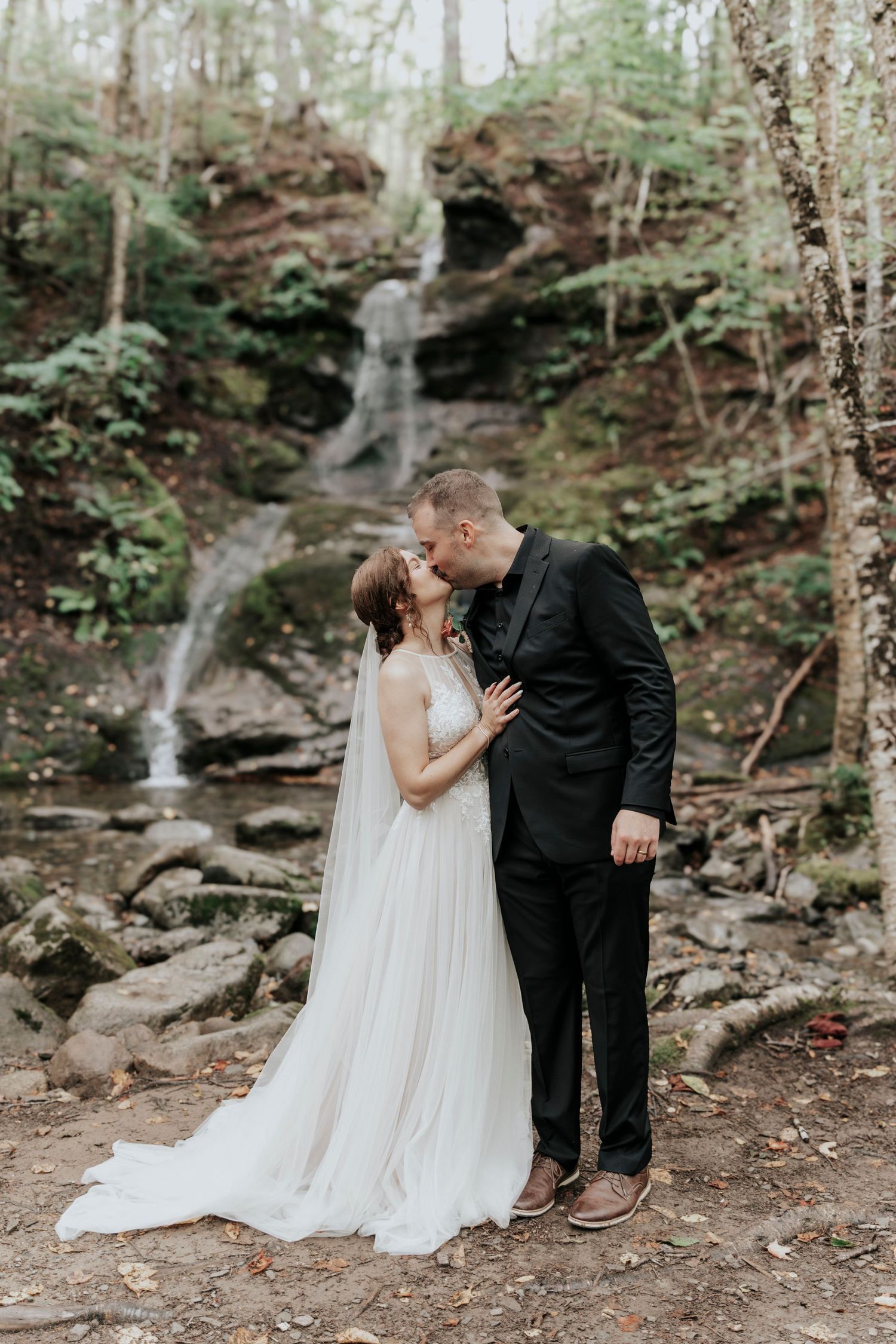 Bride and groom sharing a romantic kiss in front of a scenic waterfall surrounded by green forest.