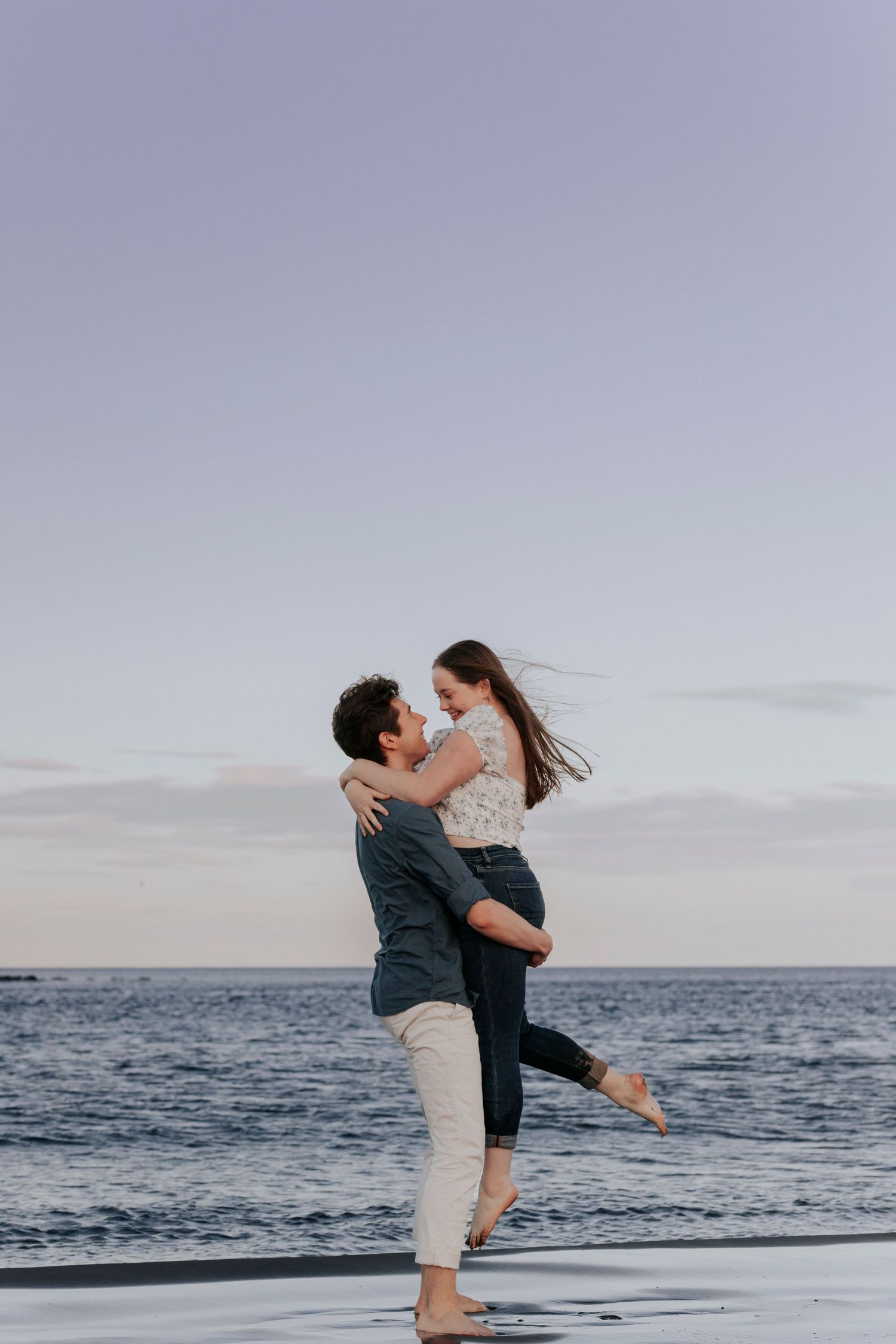 Romantic couple embracing on a beach at dusk with the man lifting the woman near the ocean.