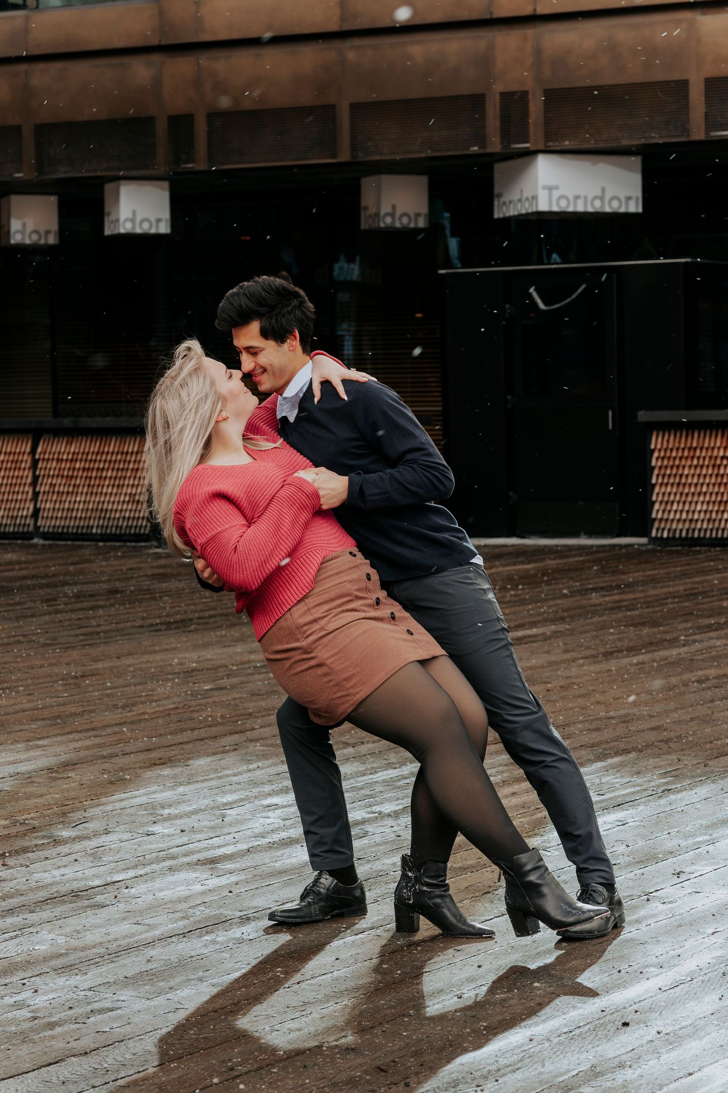 Couple performing a dramatic dance dip outside a venue, woman in red sweater and brown skirt.
