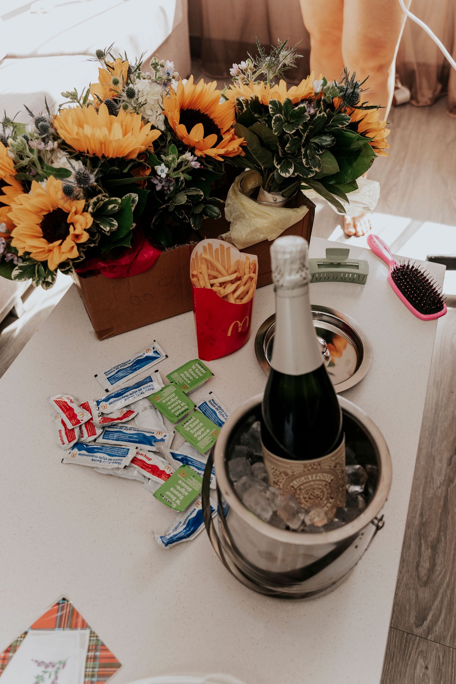 Wedding getting-ready table with sunflowers, champagne in ice bucket, McDonald's fries, and assorted accessories.