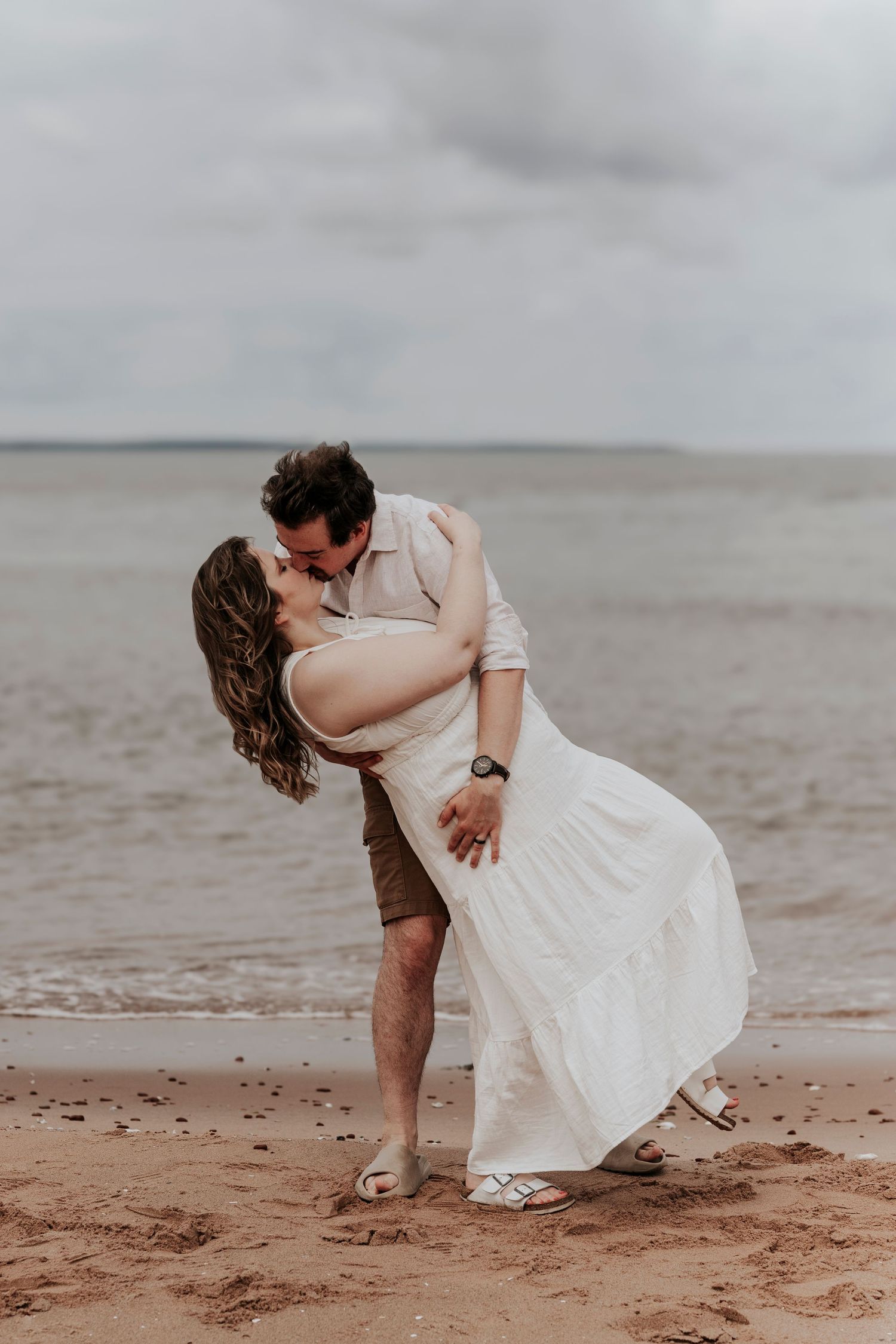 Couple sharing passionate dip kiss on sandy beach with overcast sky and ocean waves behind them.