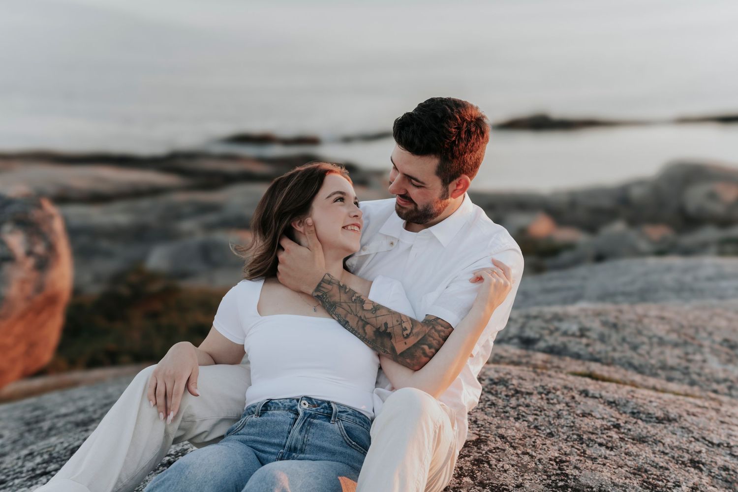 Couple embracing on rocky shore at sunset, woman in white top and jeans, man with tattoos.