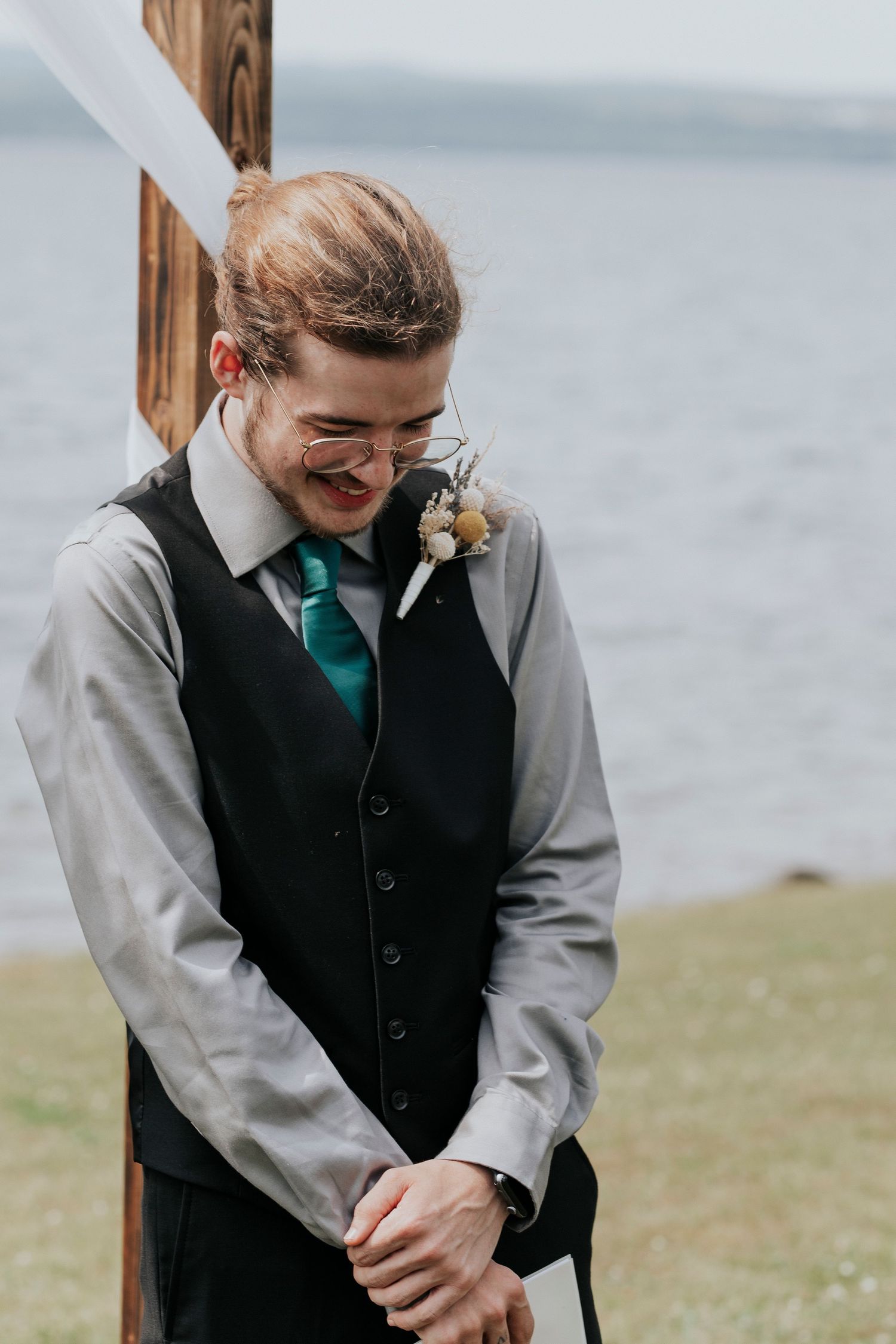 A person in a grey suit vest and emerald green tie stands smiling by the coast on a cloudy day.