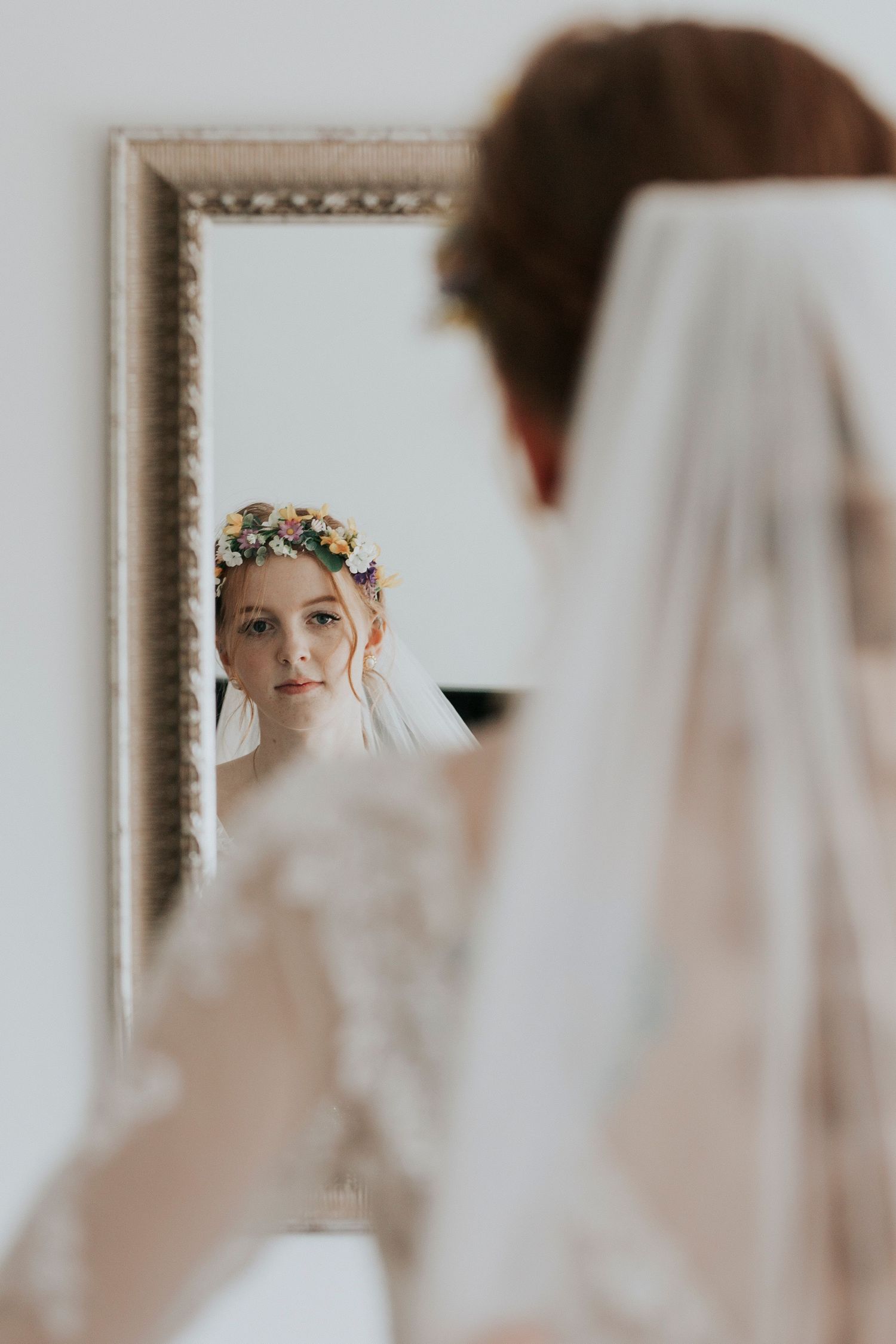 A bride wearing a floral crown and veil looks at her reflection in an ornate framed mirror.