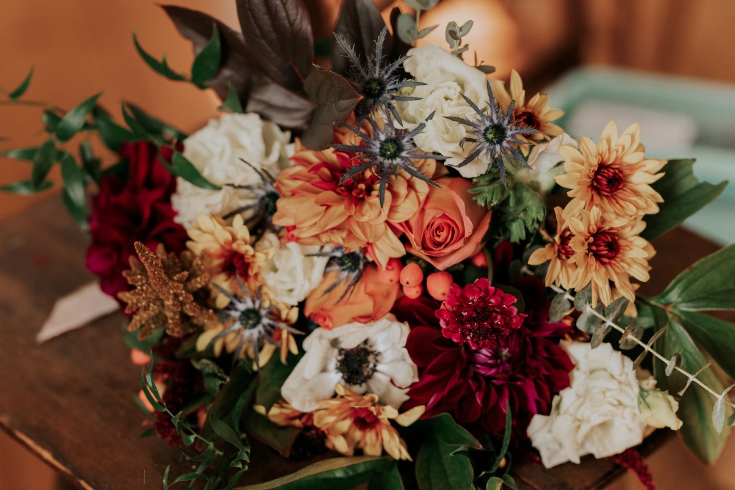 Beautiful autumn wedding bouquet featuring peach roses, deep red dahlias, white anemones and mixed greenery on wooden table.