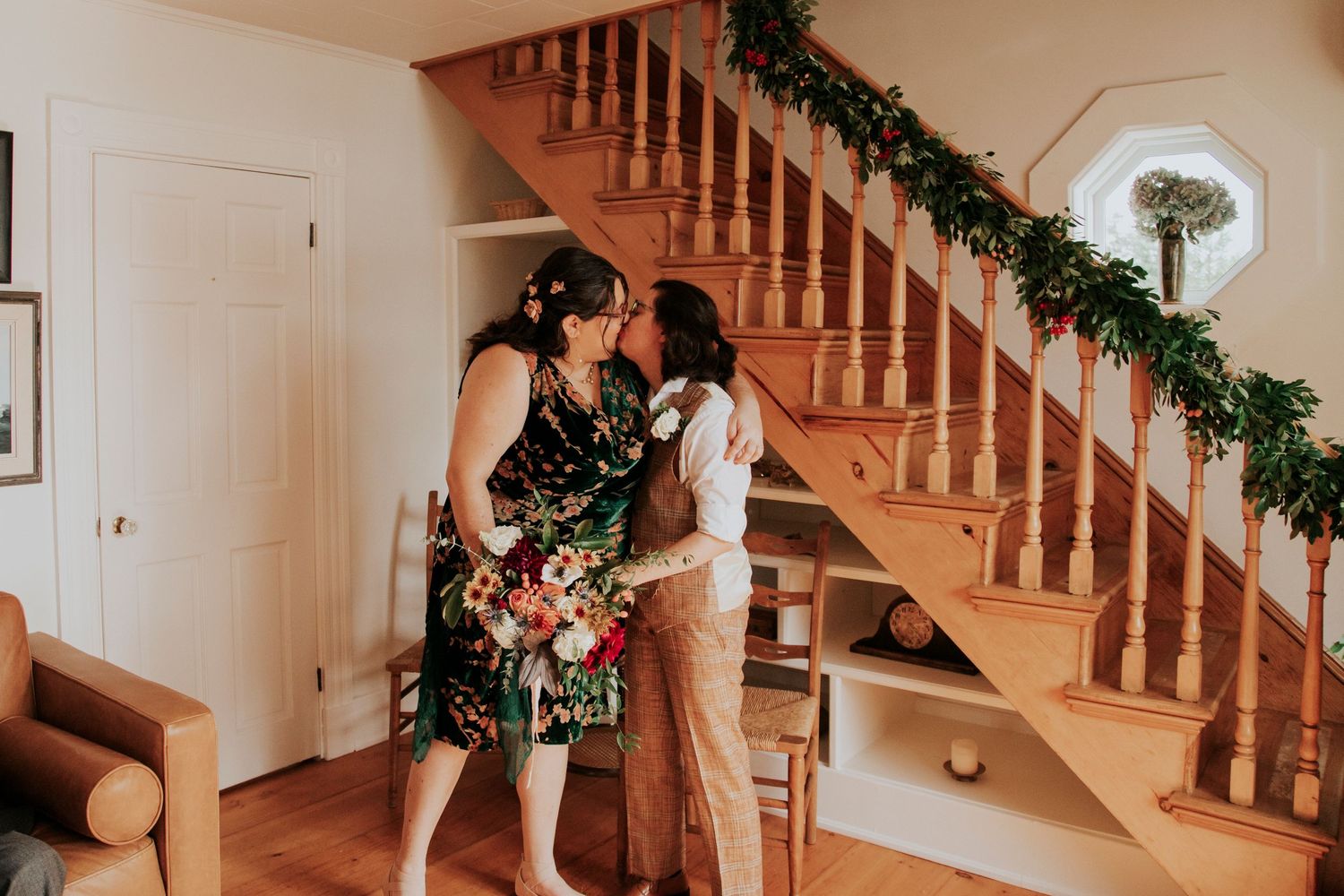 Elegant couple embraces by a wooden staircase decorated with holiday greenery and floral arrangements.