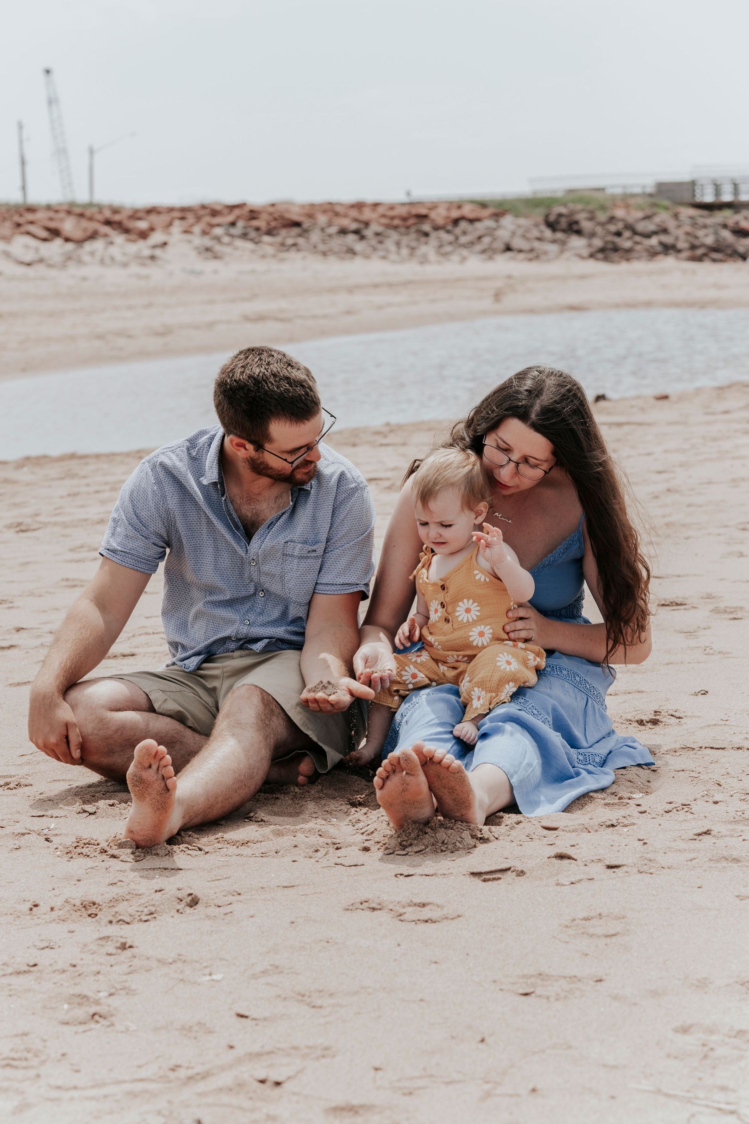 Young family sitting on sandy beach examining shells with their baby girl in yellow floral romper.