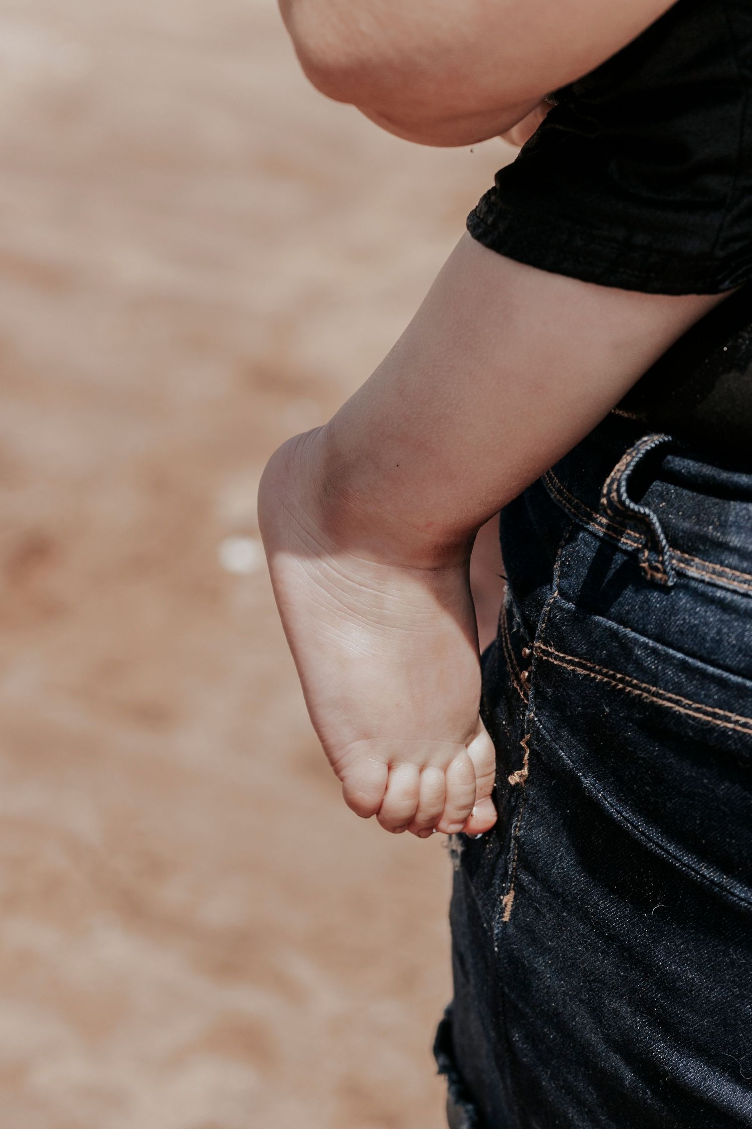 Close-up of a baby's bare foot resting against adult denim jeans on a sandy beach.