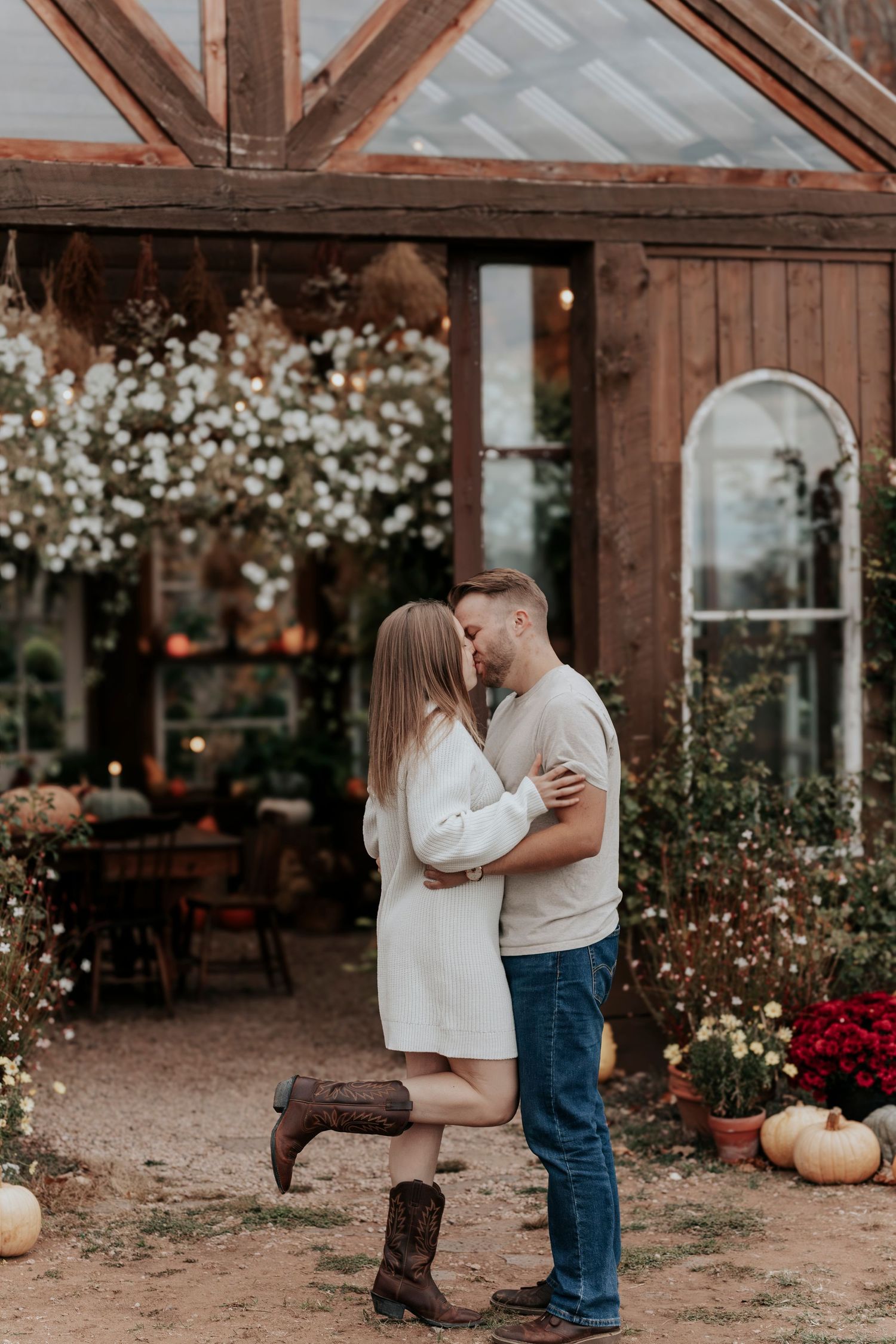 Couple kissing outside a rustic wooden greenhouse surrounded by autumn flowers and pumpkins.