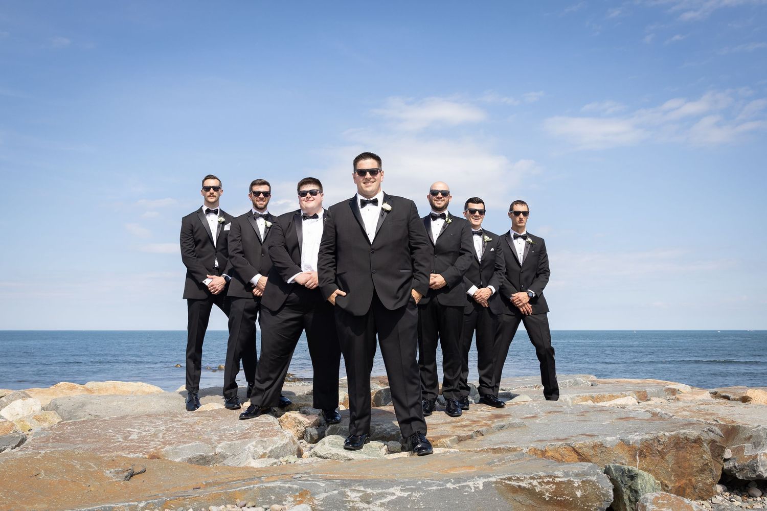 Groom and groomsmen in black tuxedos and bow ties posing on rocky oceanside cliffs under a clear blue sky.
