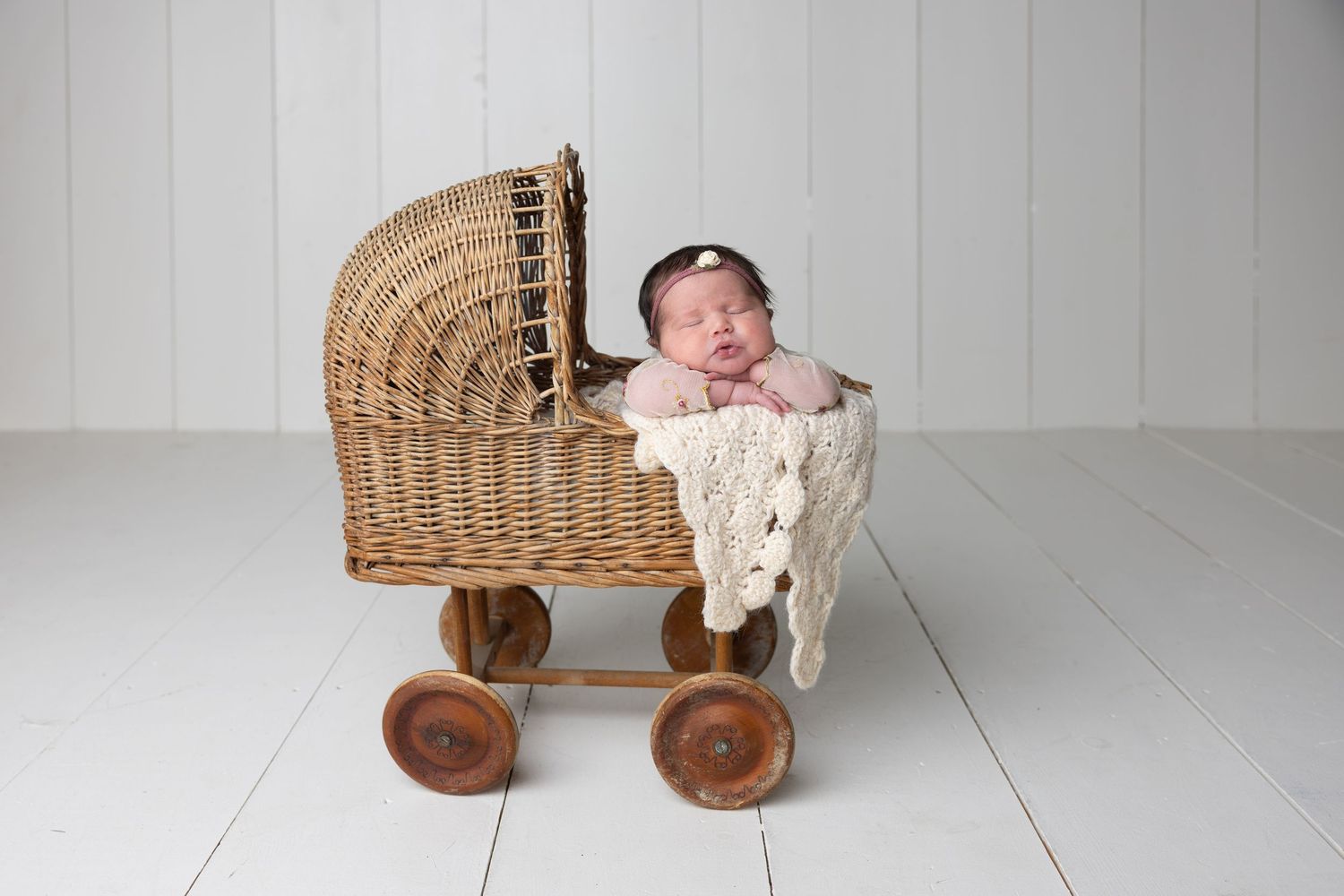 Newborn baby sleeping peacefully in a vintage wicker pram with wooden wheels on a white floor.
