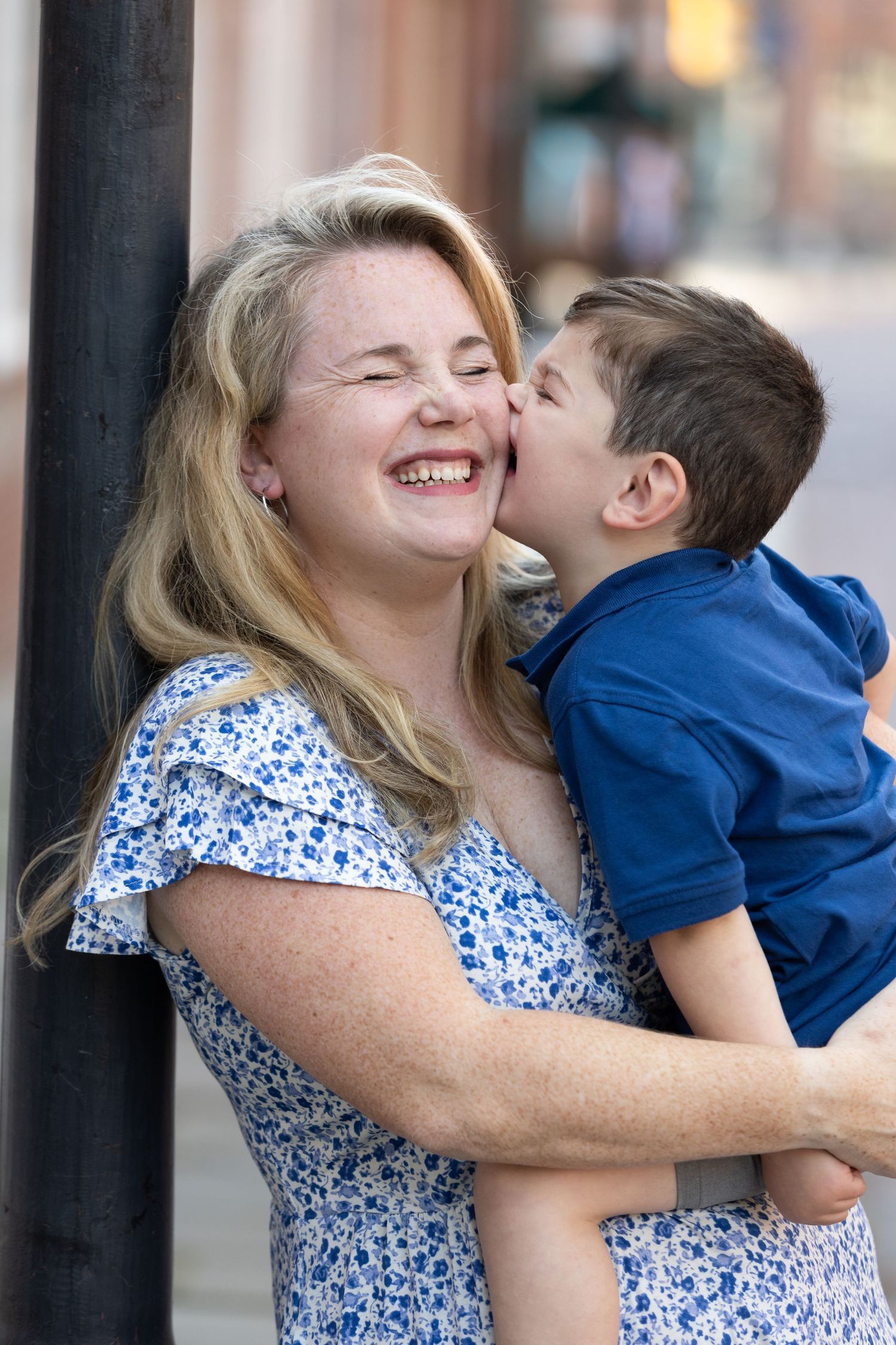 A laughing blonde woman in a floral dress holds a young boy in a blue shirt who kisses her cheek outdoors.