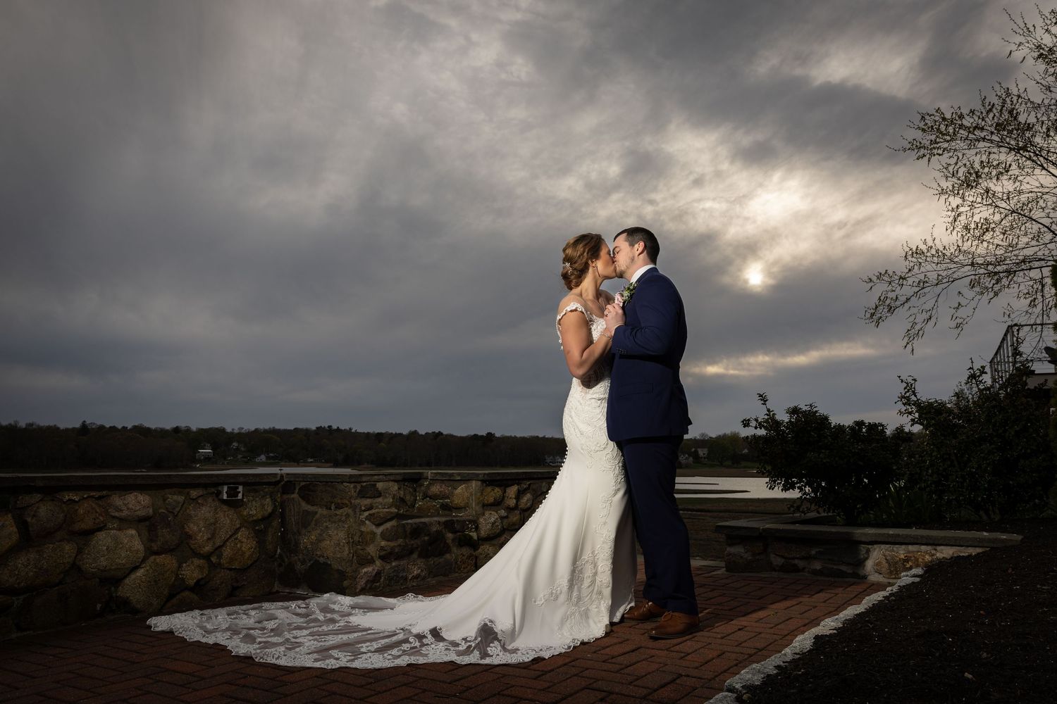 Bride and groom share a romantic kiss outdoors under dramatic stormy skies at their elegant wedding venue.