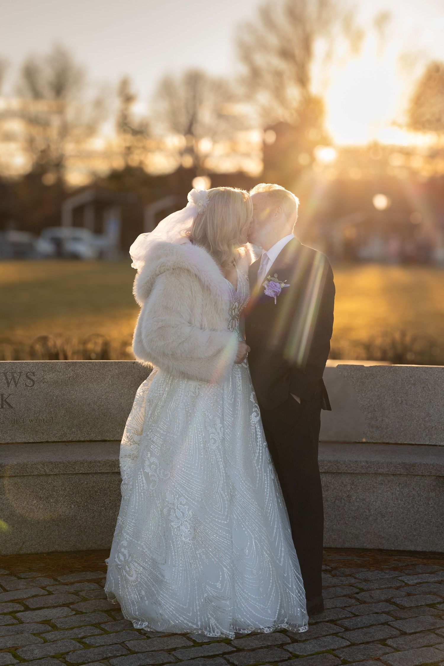 Bride and groom sharing a romantic sunset kiss, bride wearing white lace gown and fur wrap outdoors.