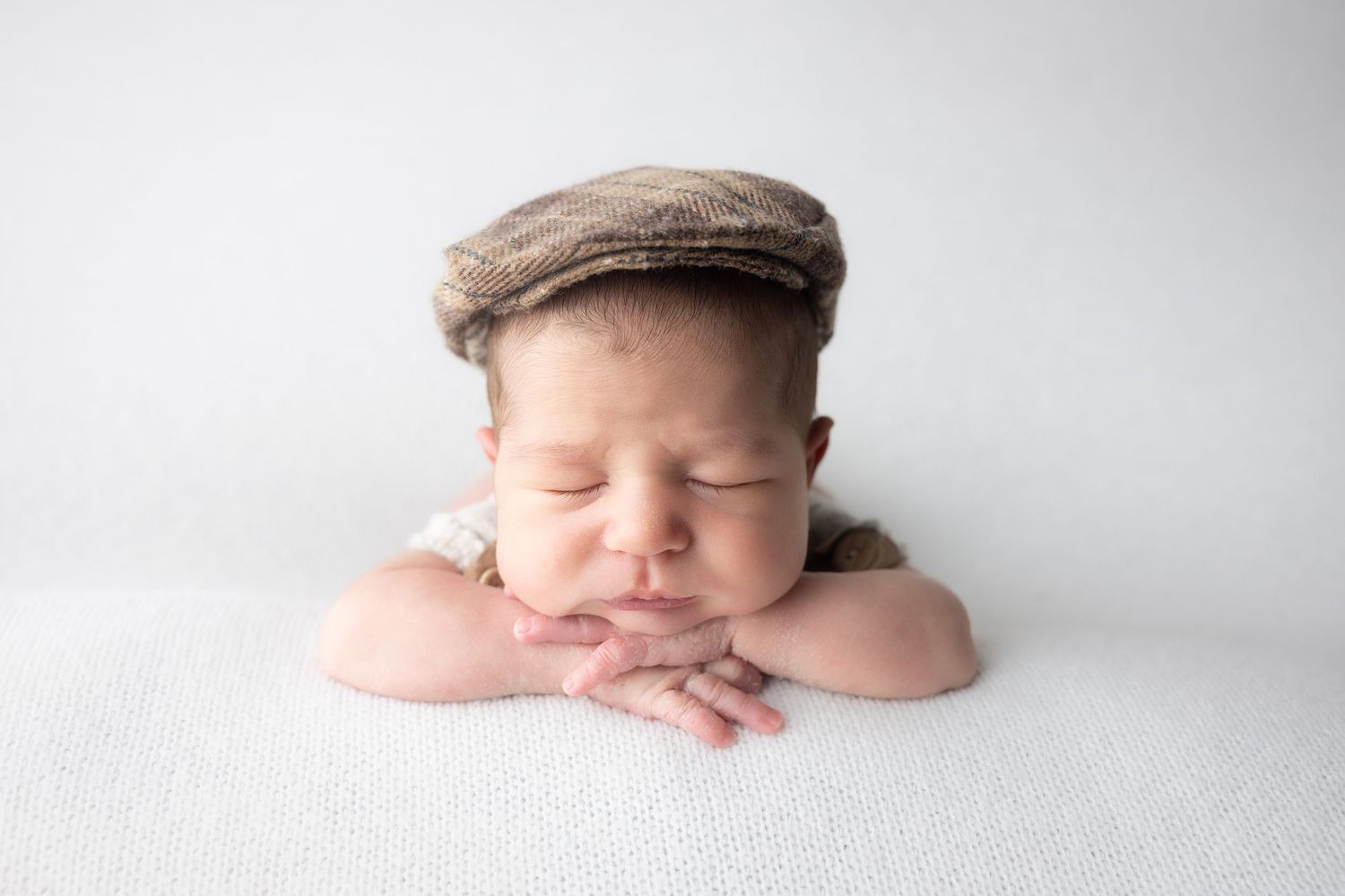 Sleeping newborn baby wearing a brown newsboy cap, resting chin on folded hands on a white surface.