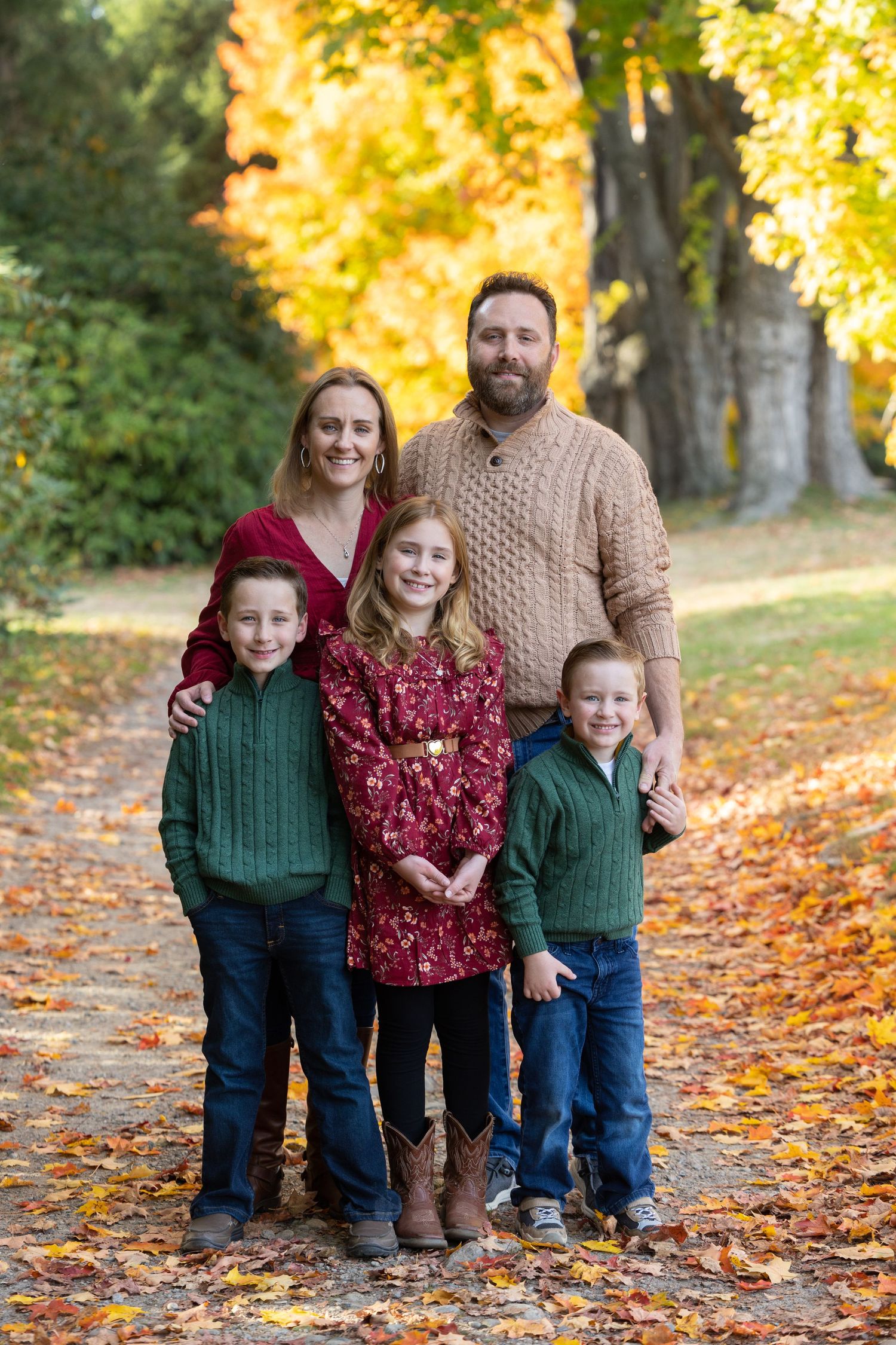 Family of five posing outdoors in autumn, wearing coordinated fall outfits among colorful foliage on a leaf-covered path.