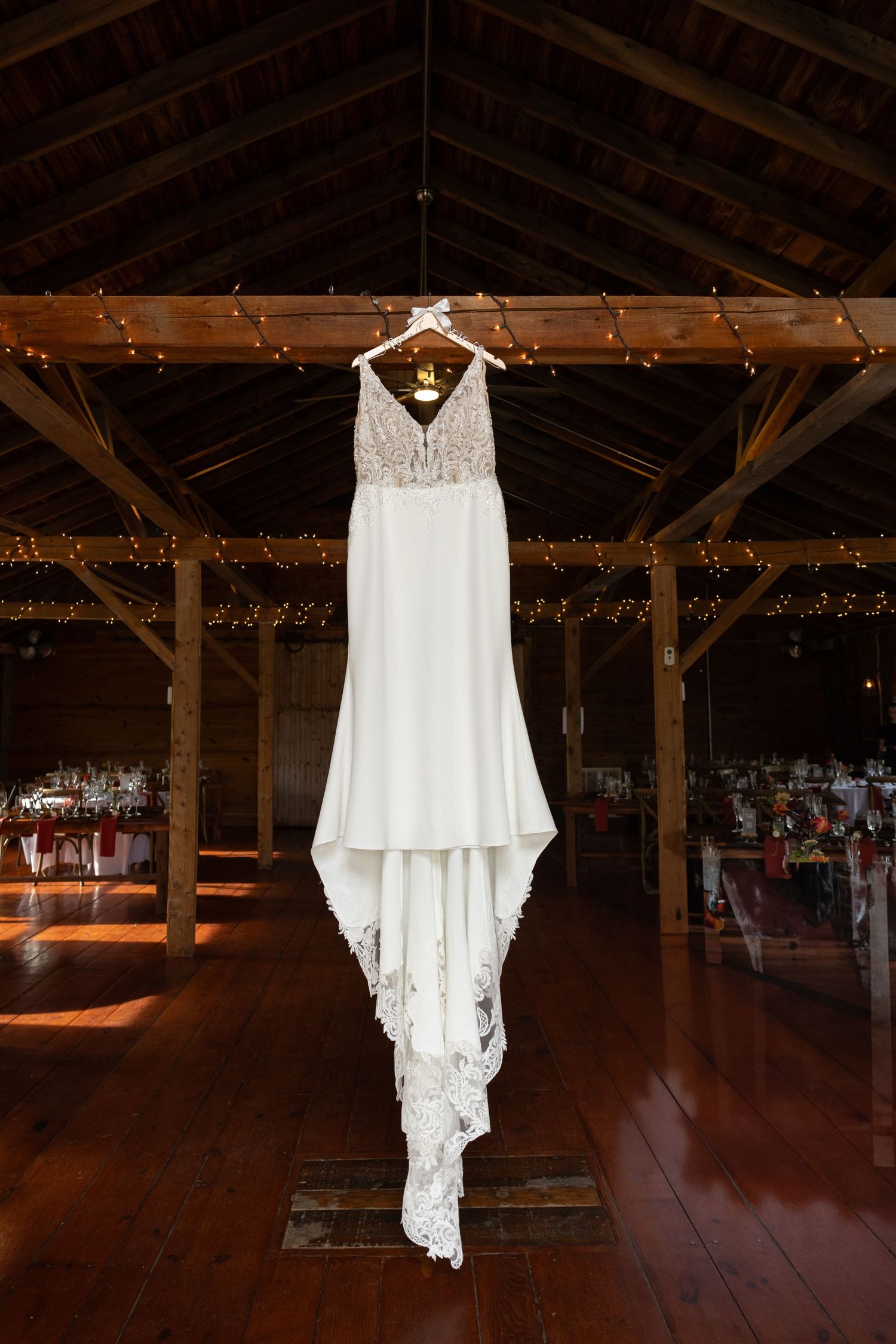 White lace wedding dress hanging from wooden beam in rustic barn venue decorated with string lights.